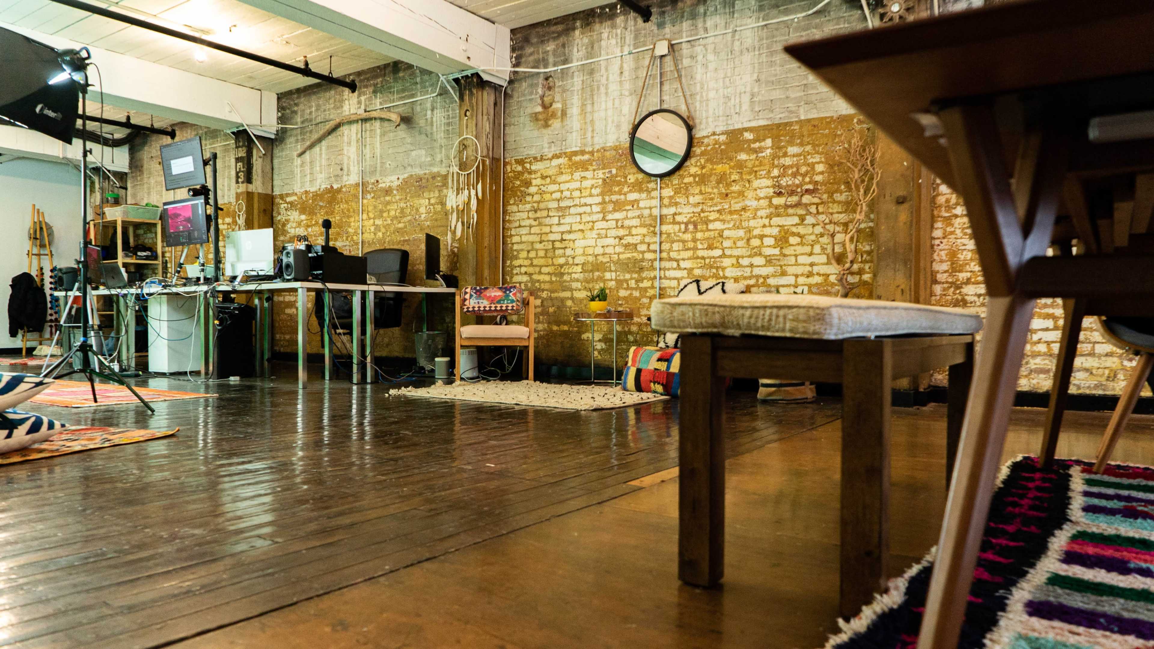The image shows an industrial-style workspace with wooden flooring, exposed brick walls, and various furniture items, including a bench and a desk setup.