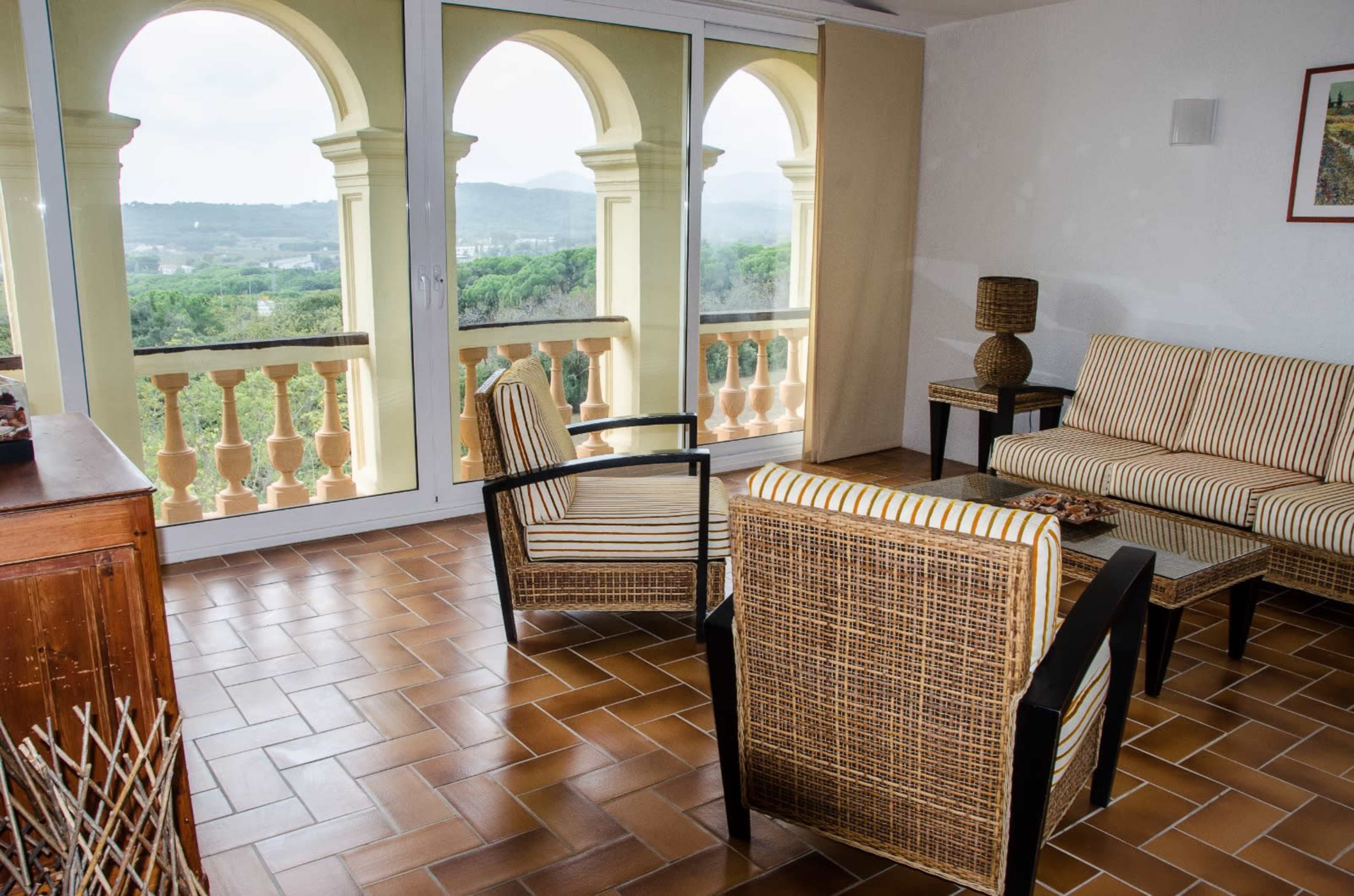 The room features a seating area with two wicker chairs and a sofa, set against large glass doors that open to a balcony with a view of a green landscape.