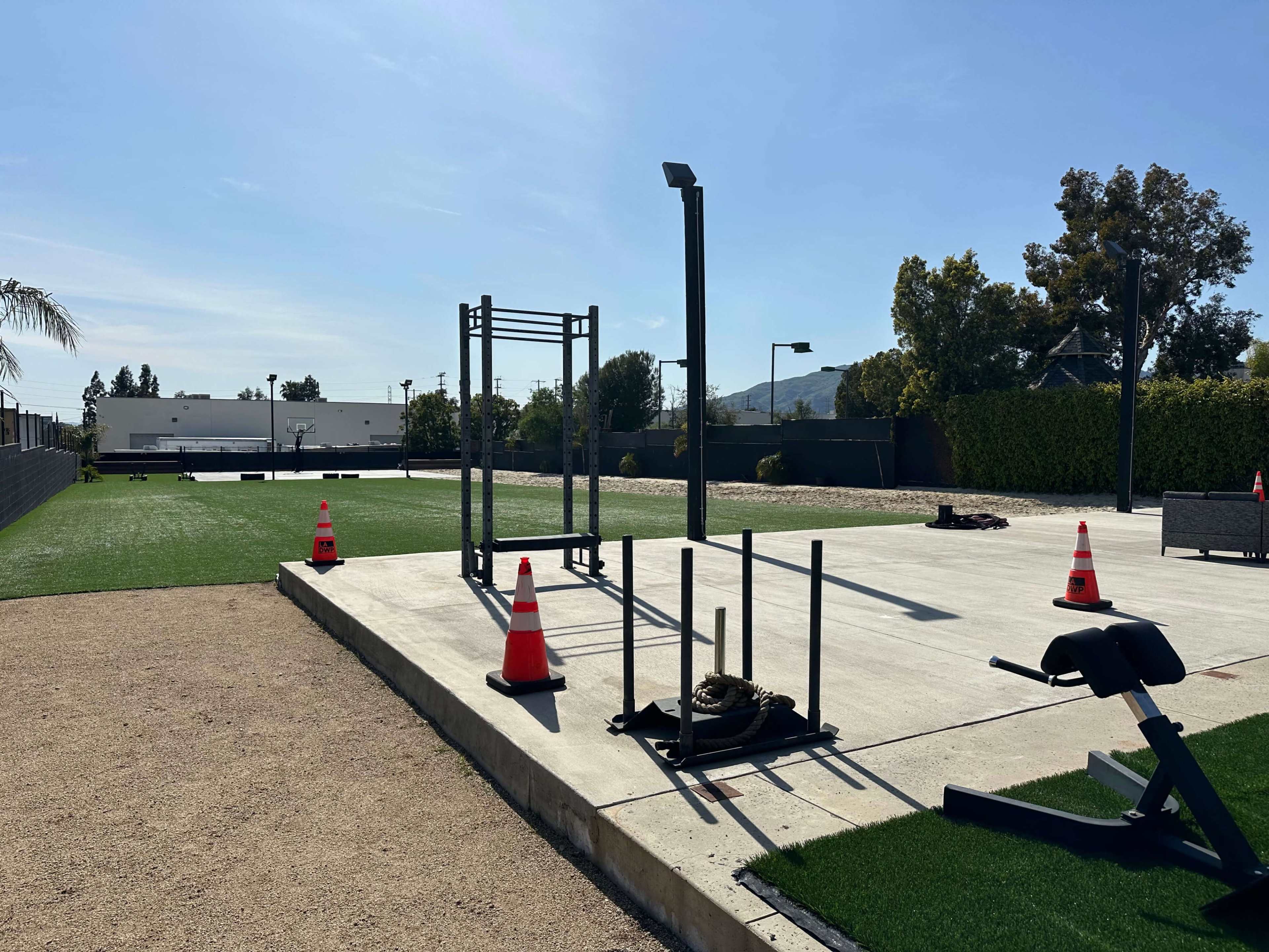 The image shows an outdoor gym area with workout equipment and traffic cones on a concrete surface, surrounded by green grass and trees.