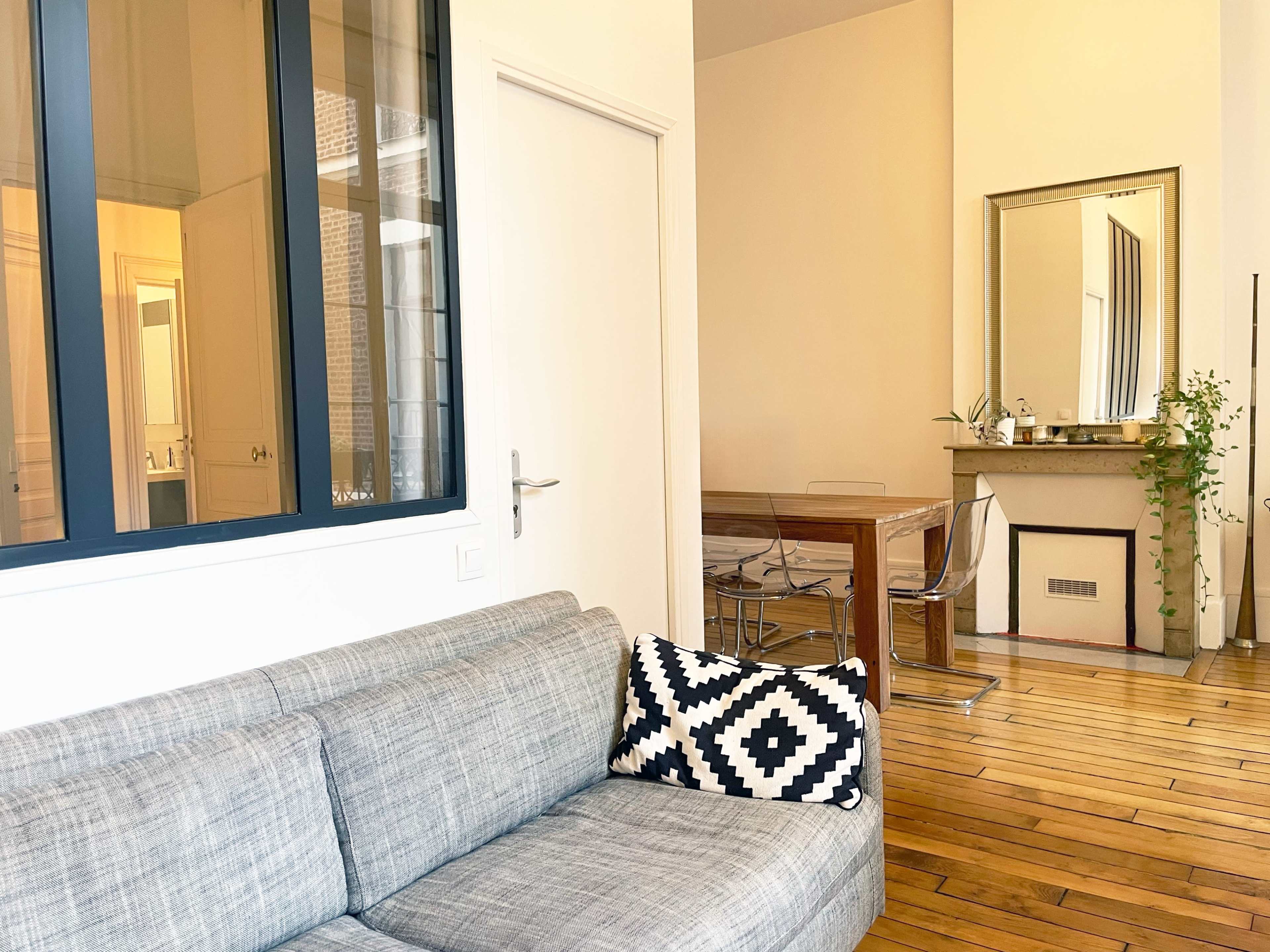 The image shows a living room with a gray sofa, a patterned cushion, a dining area featuring a wooden table, and a mirror on the wall.