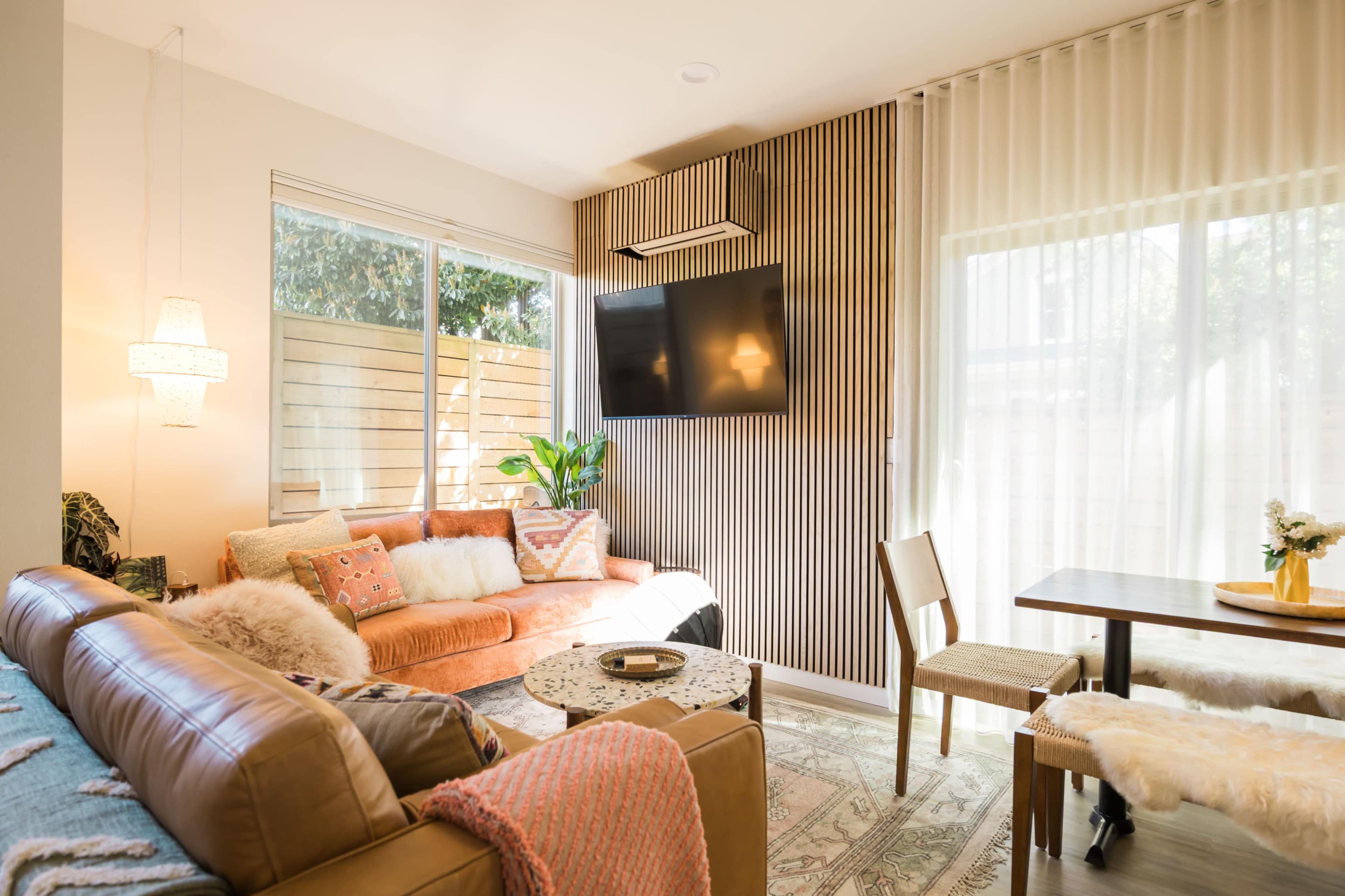 The image shows a cozy living room with a sectional sofa, a flat-screen TV mounted on the wall, and large windows allowing natural light to fill the space.