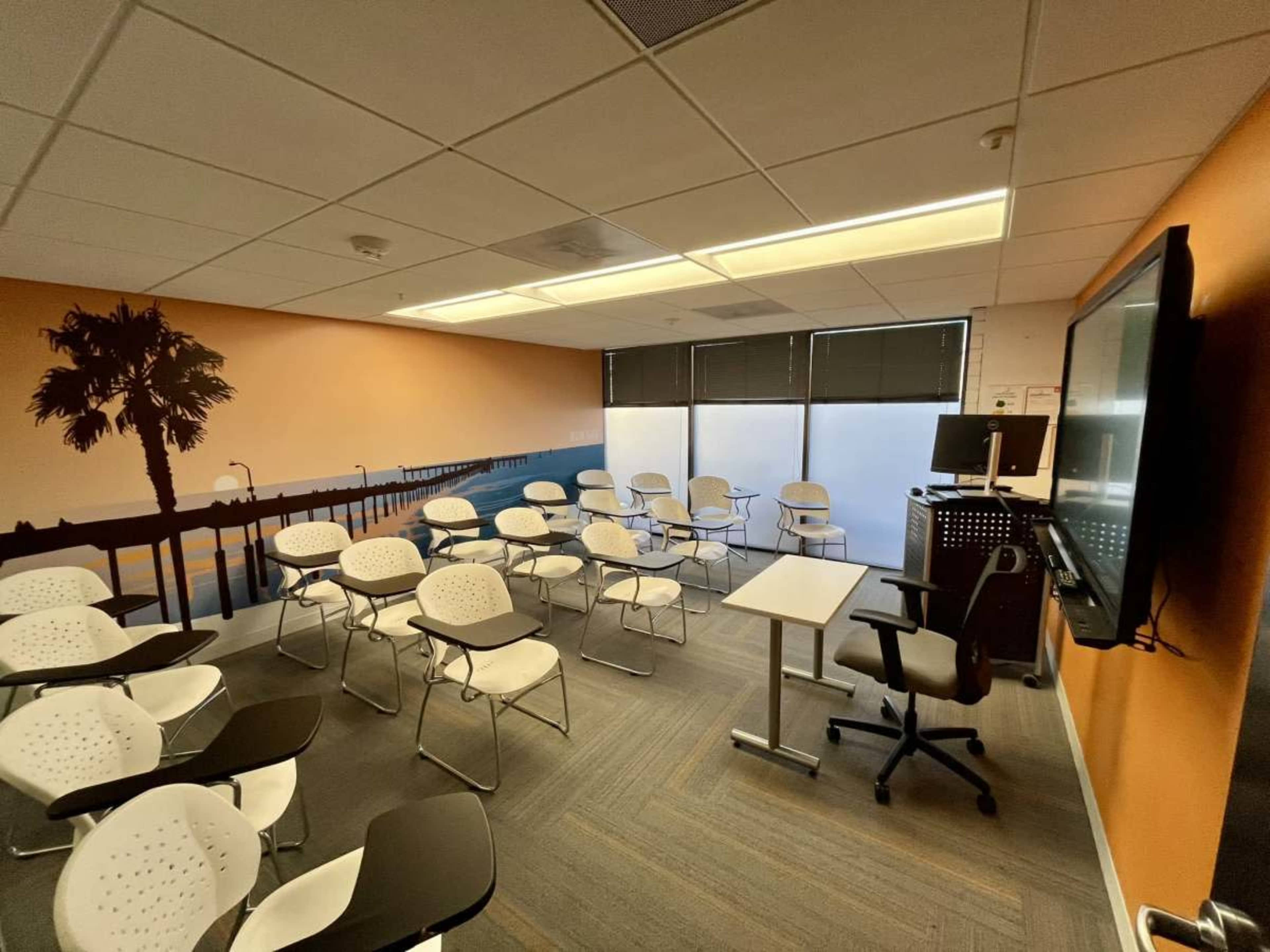A classroom with white chairs arranged in rows, a large screen on the front wall, and a mural featuring a palm tree and ocean scene.
