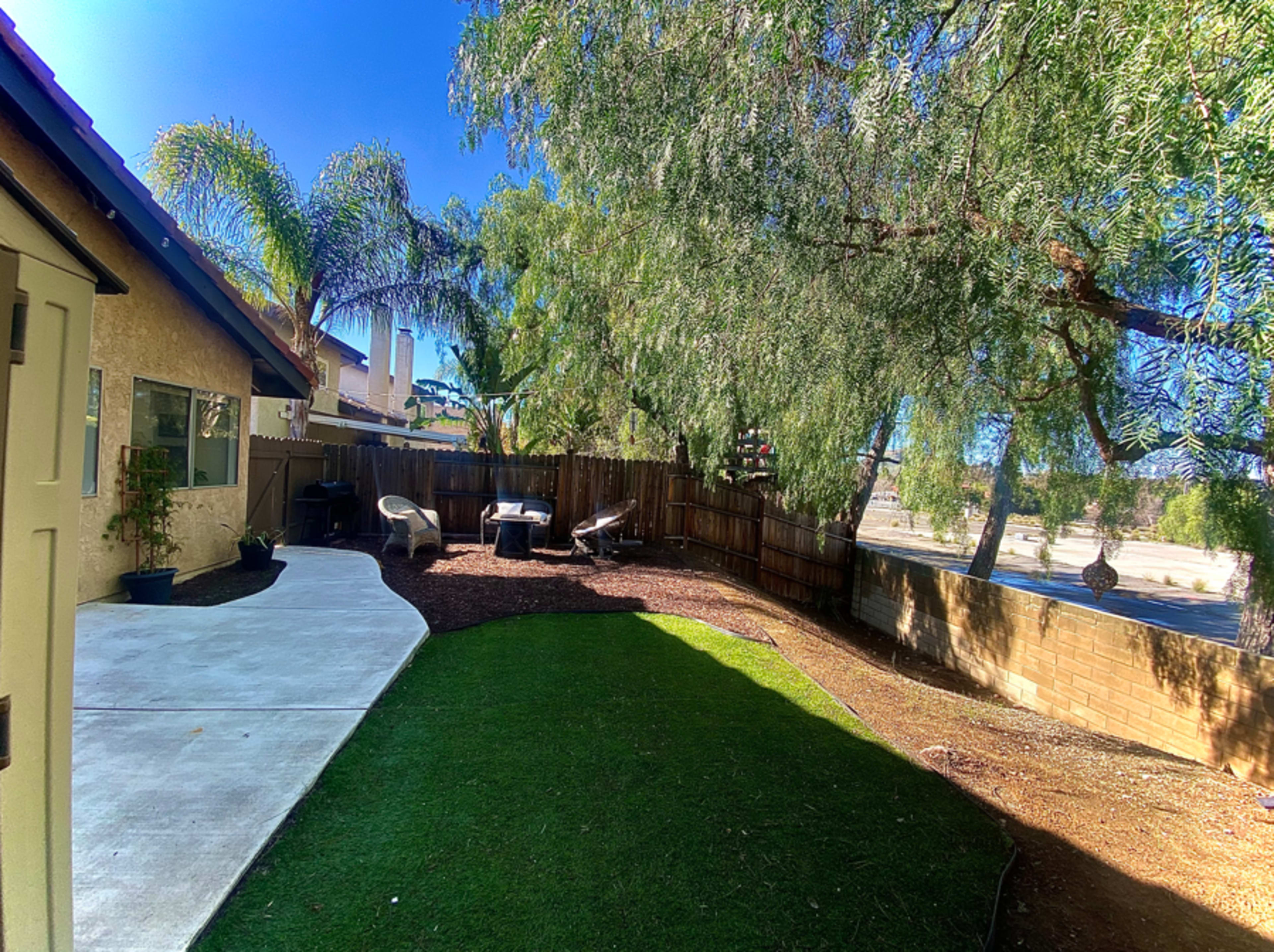 A well-maintained backyard features a small grassy area surrounded by trees, with a concrete walkway leading to a seating area with chairs.