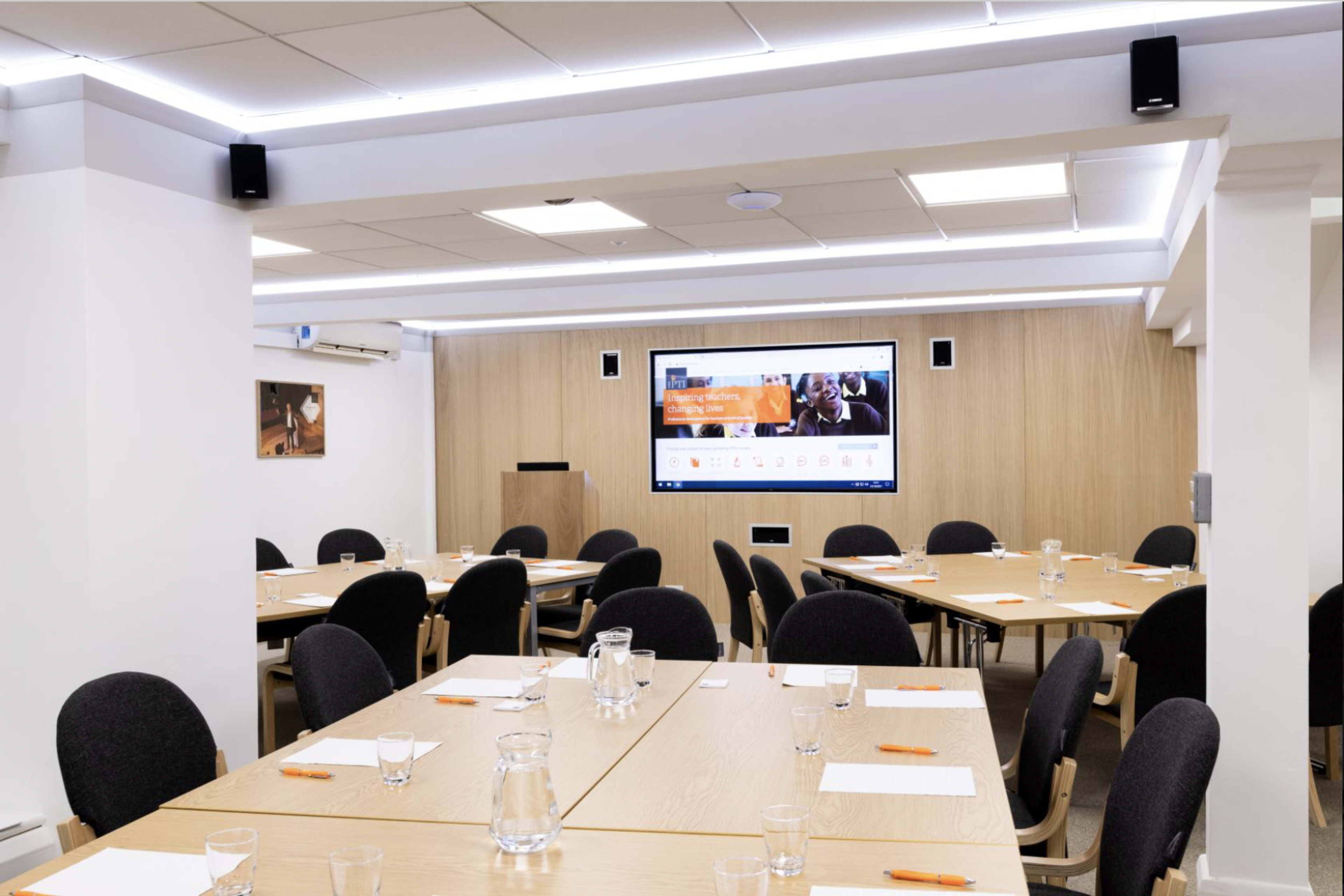 A conference room is set up with multiple tables, chairs, and a screen displaying information on a wall.