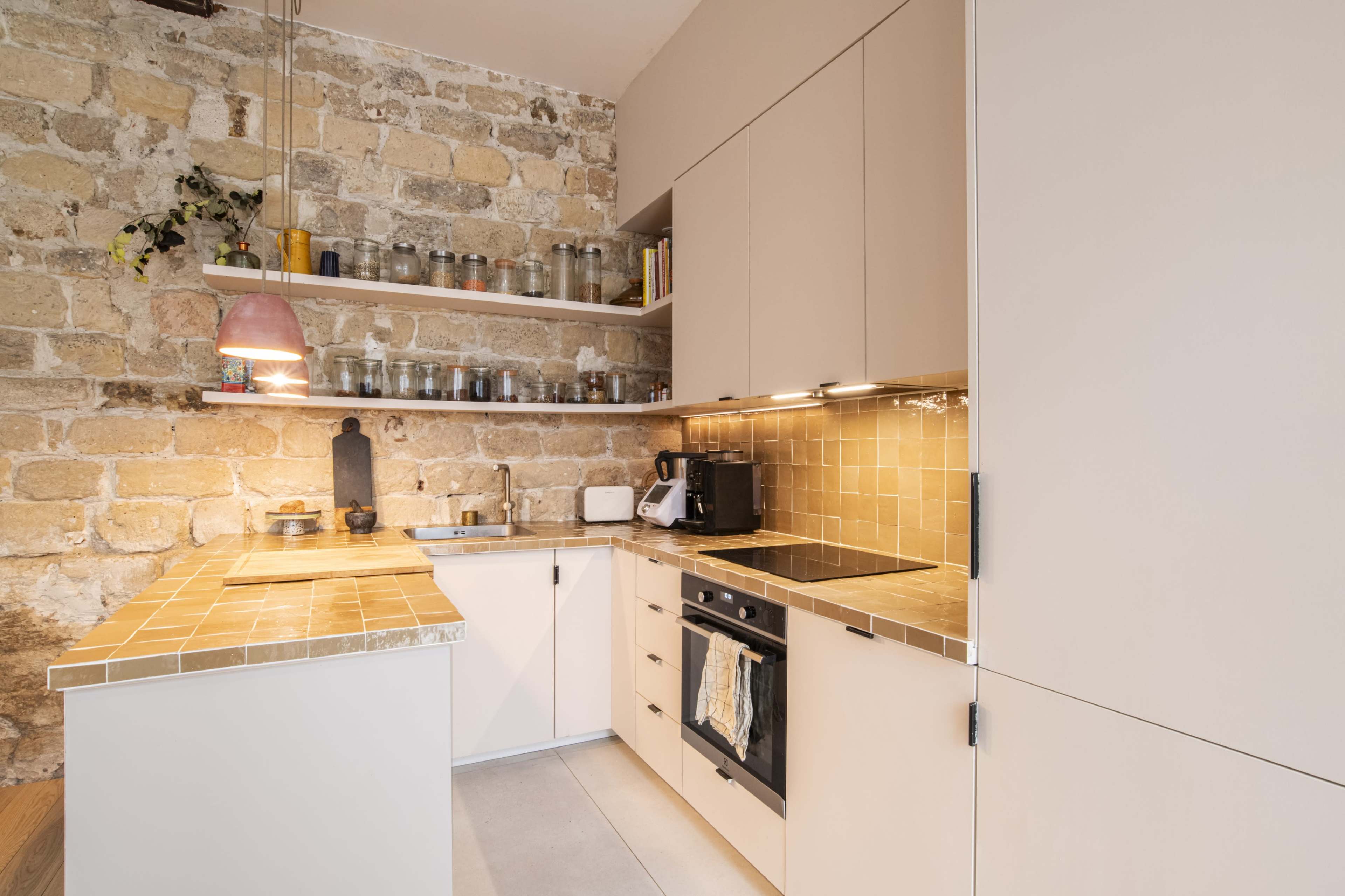 The image shows a modern kitchen with beige cabinetry, a stone wall, and a countertop featuring square tiles.