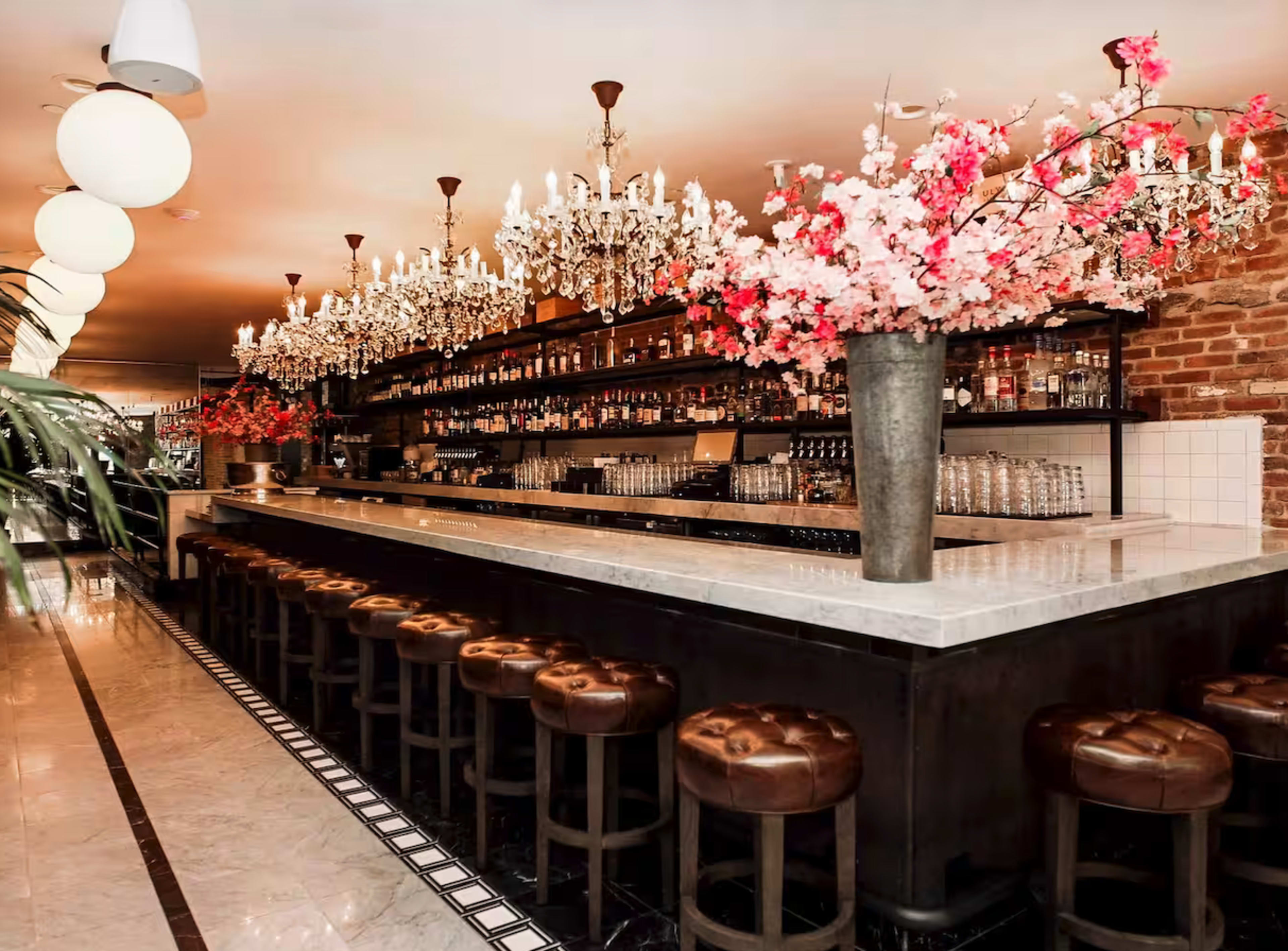 The image shows a stylish bar with a long white countertop, leather-stooled seating, and chandeliers, adorned with pink cherry blossoms.