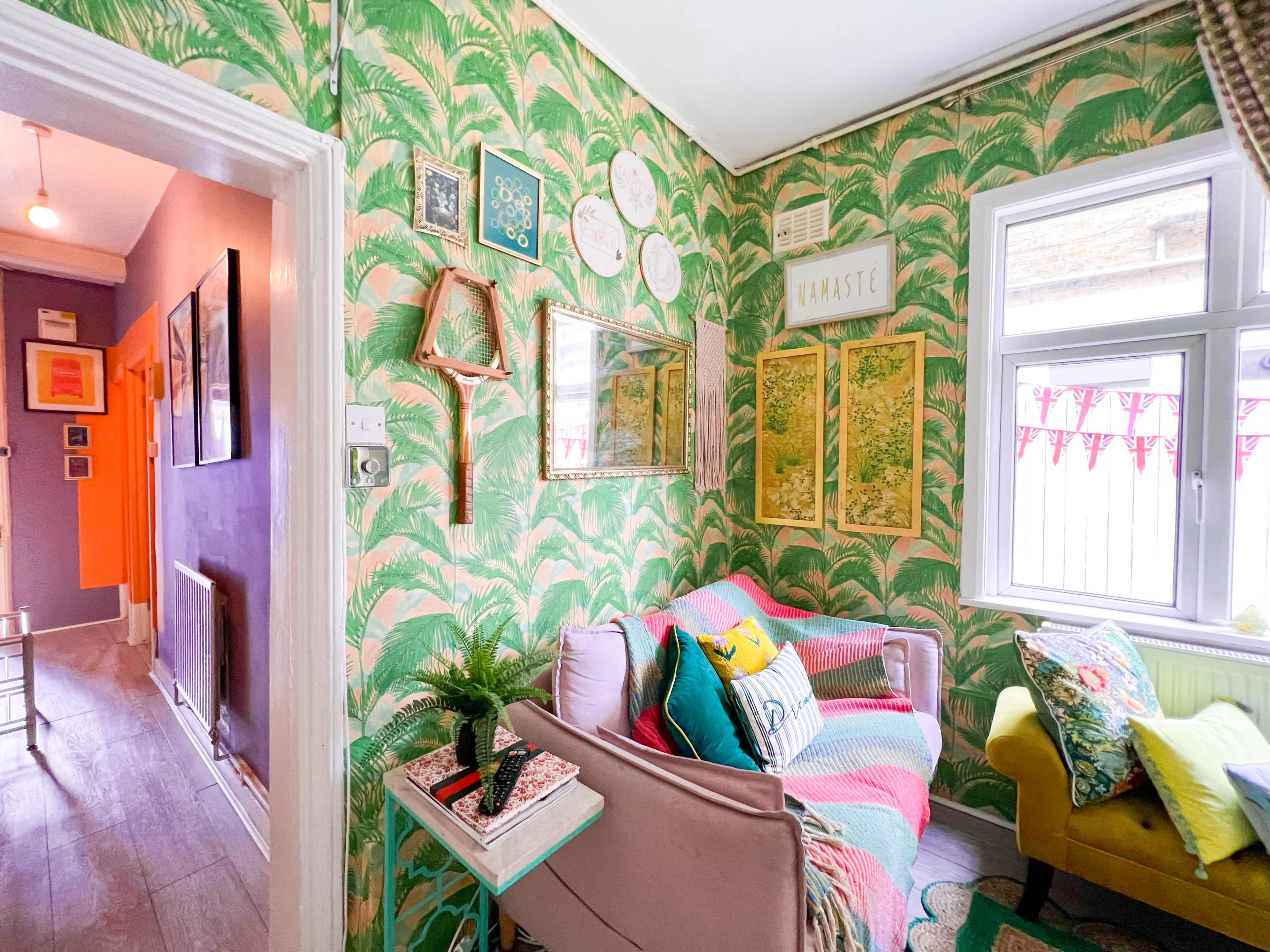 The image shows a vibrant living room with patterned wallpaper, a couch adorned with colorful cushions, and framed artworks on the walls.