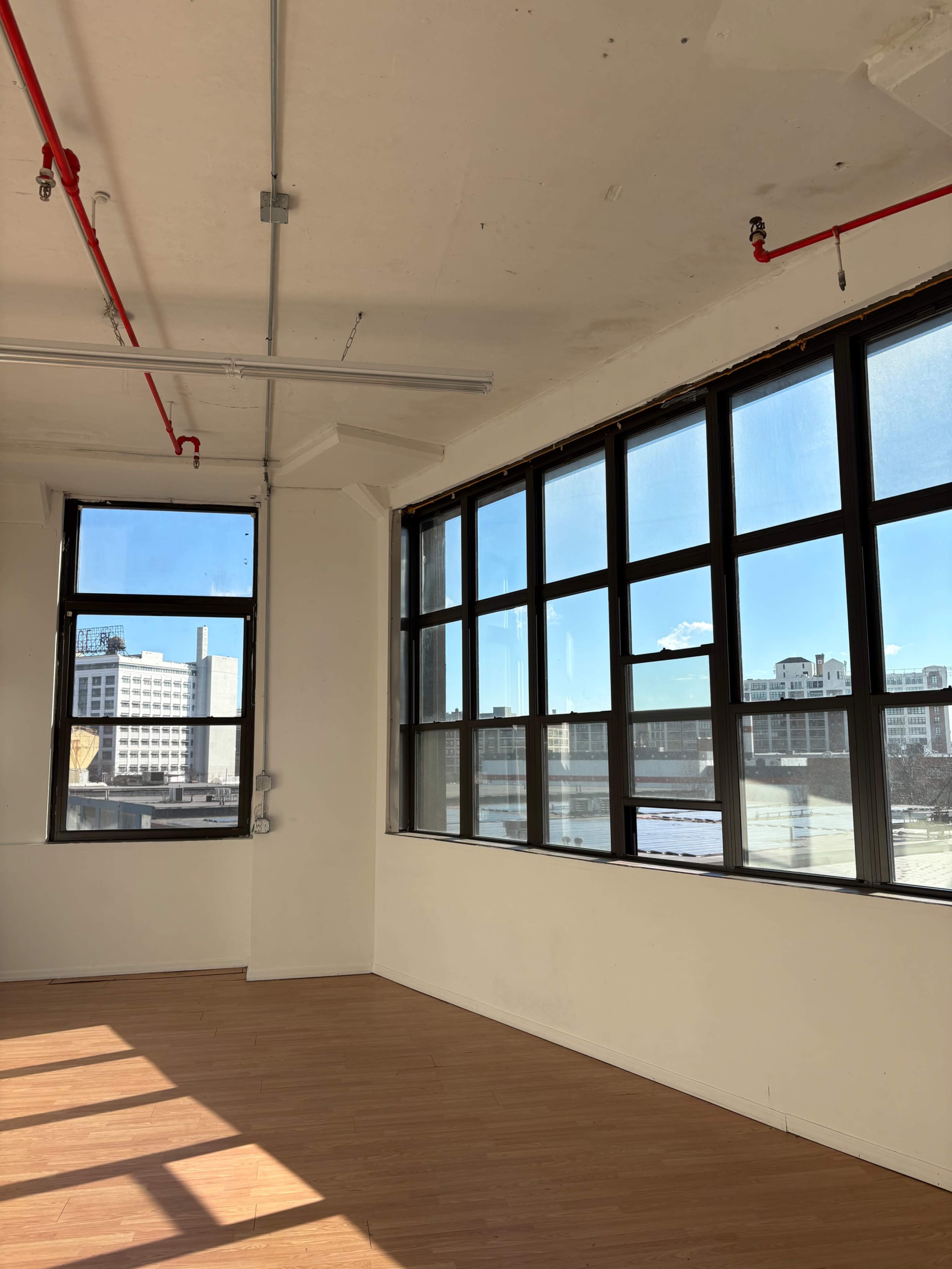 The image shows a bright, empty room with large windows and wooden flooring, allowing sunlight to fill the space.