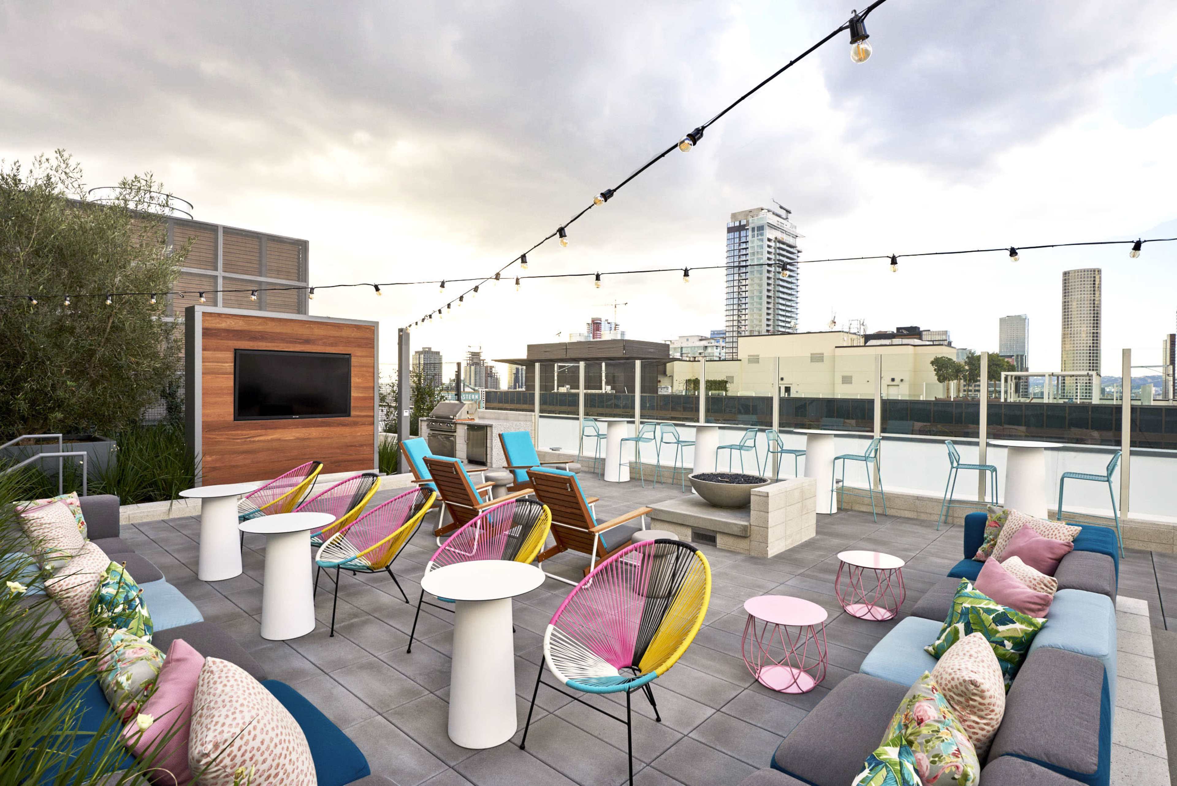A rooftop terrace with colorful chairs, tables, and a fire pit, surrounded by a city skyline and string lights overhead.