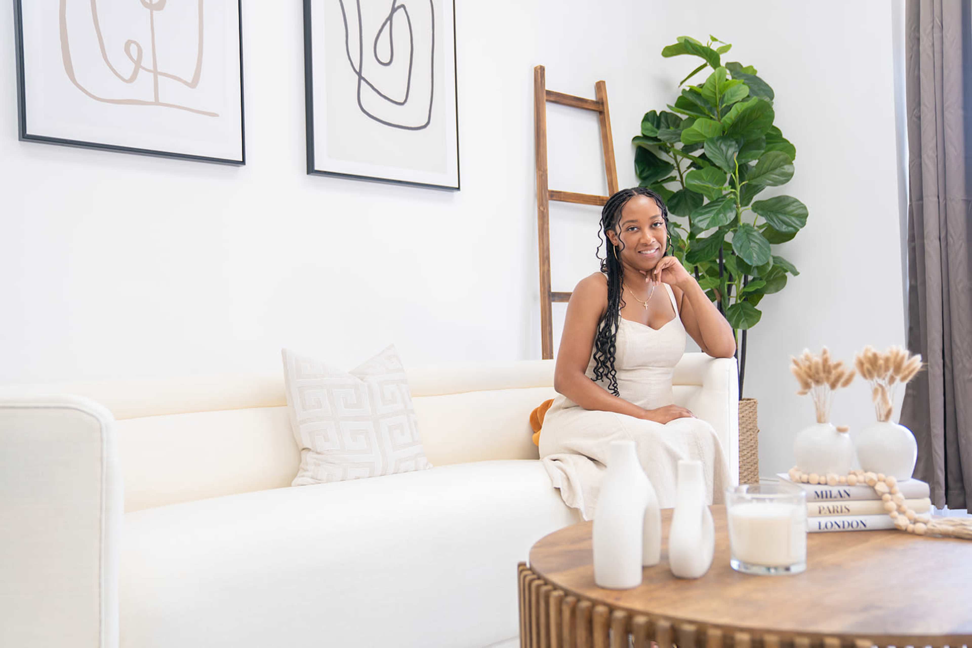 A young woman sits on a cream-colored sofa in a bright, minimalistic living room adorned with abstract wall art and indoor plants.