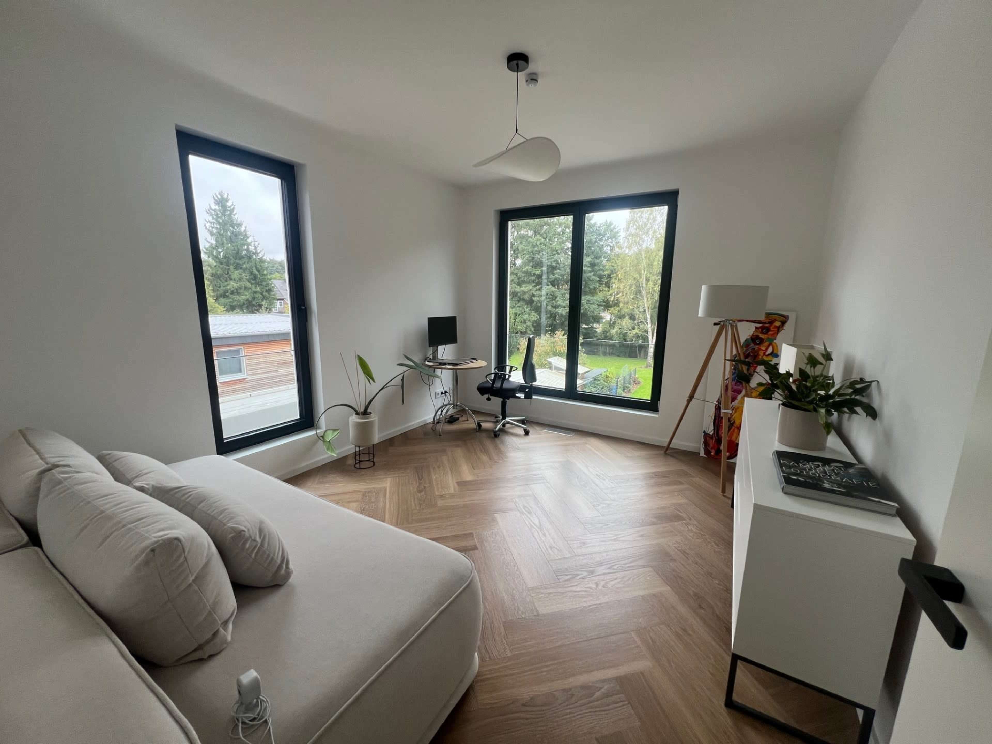 A bright room features large windows overlooking a green landscape, a small desk with a chair, and a modern sofa.