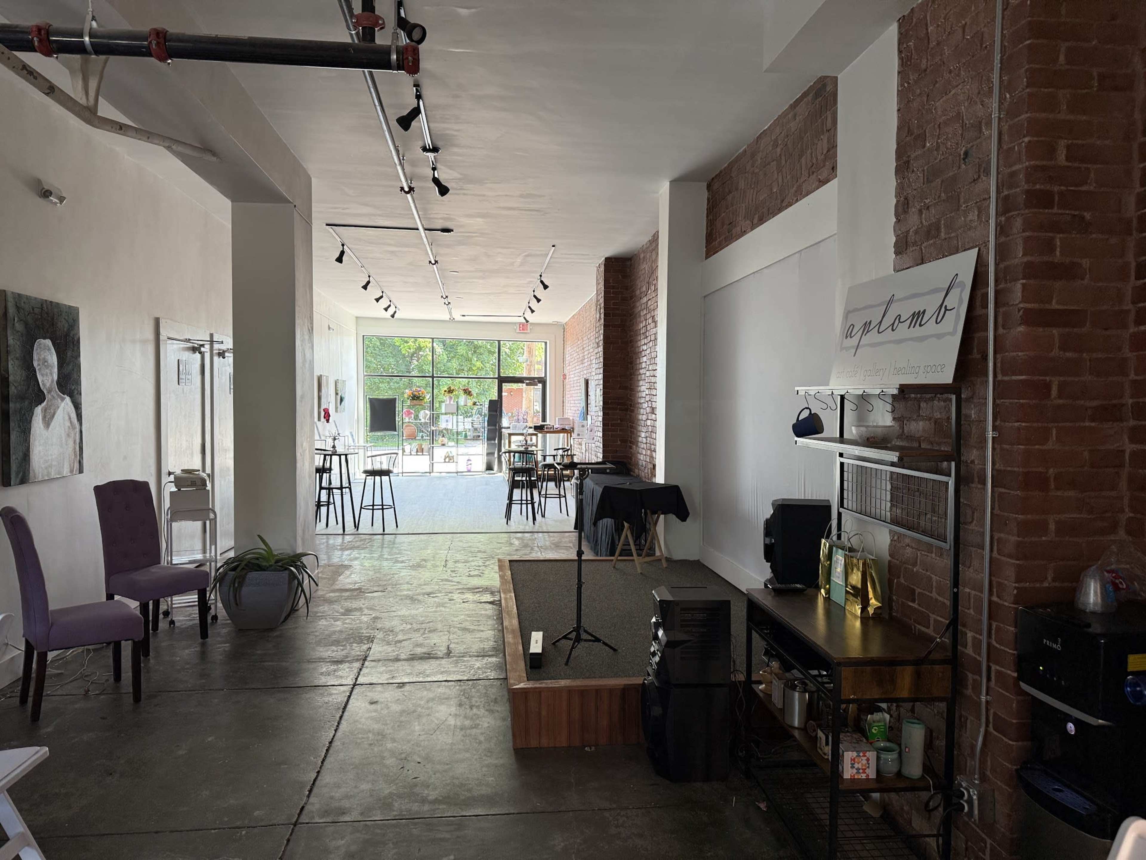 The image shows an empty interior space with exposed brick walls, tables, and chairs arranged for an event, and a small stage area with a microphone stand.