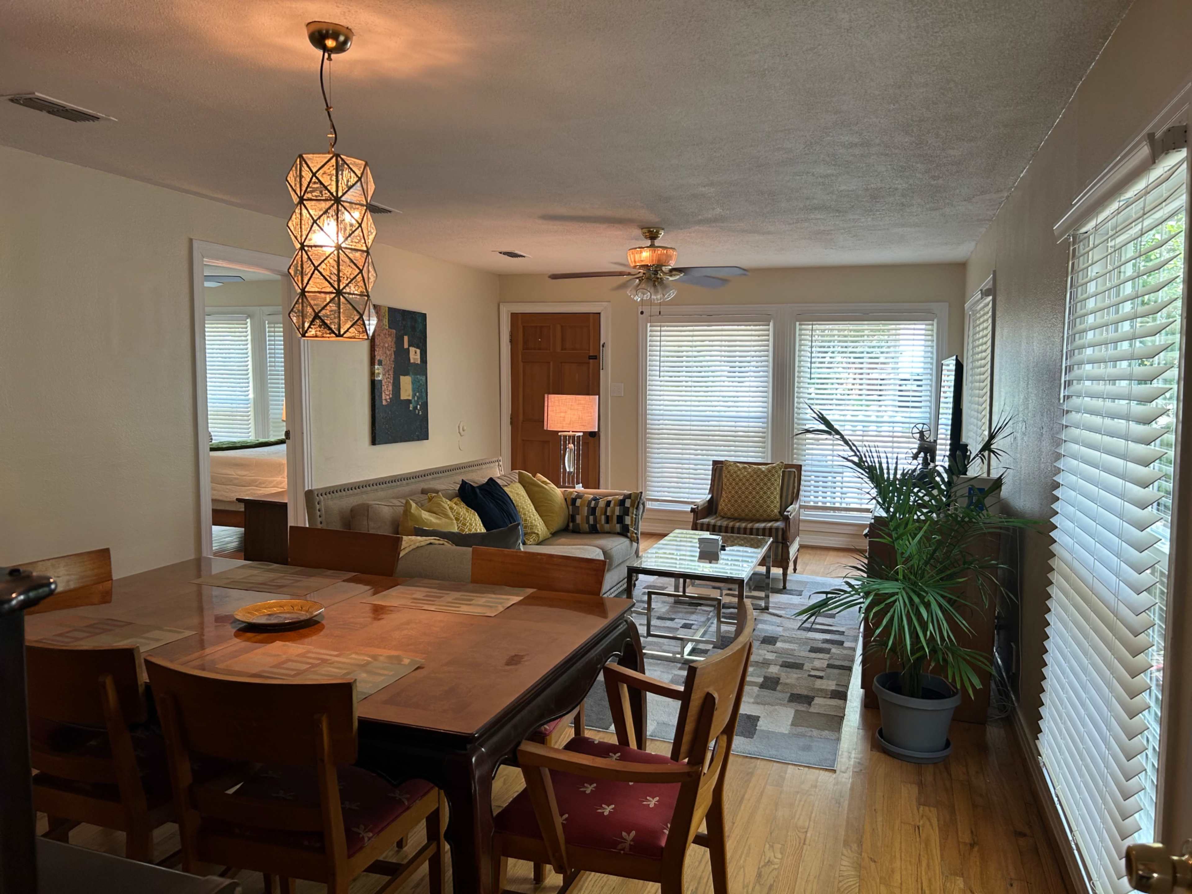 The image shows a living space with a dining area featuring a wooden table and chairs, and a seating area with a couch, armchair, and coffee table, surrounded by large windows.