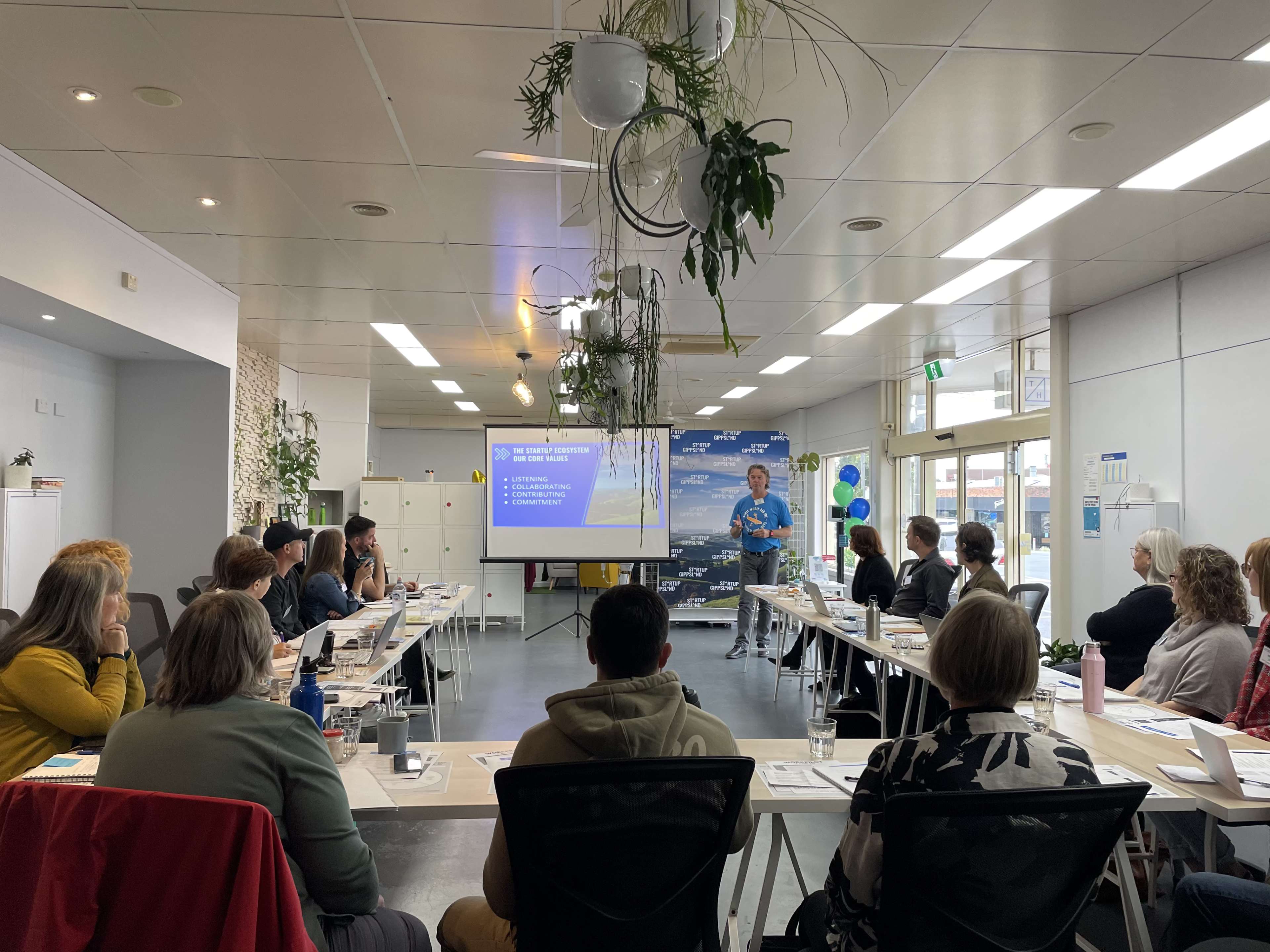 A presenter stands in front of a group seated at tables in a conference room, with a projector screen displaying information in the background.