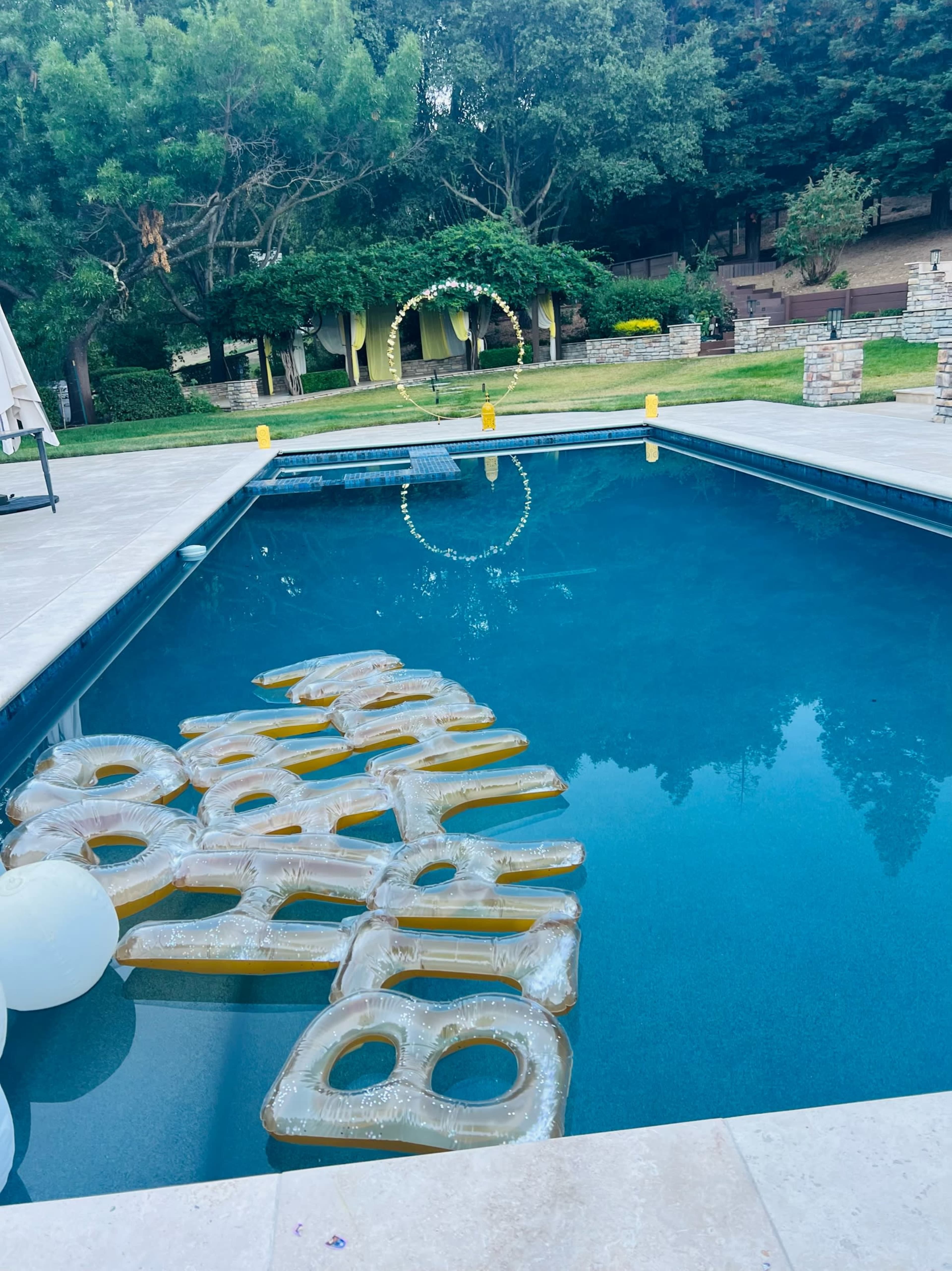 The image shows a swimming pool with oversized, transparent letters floating on the water, surrounded by greenery and a decorative arch in the background.