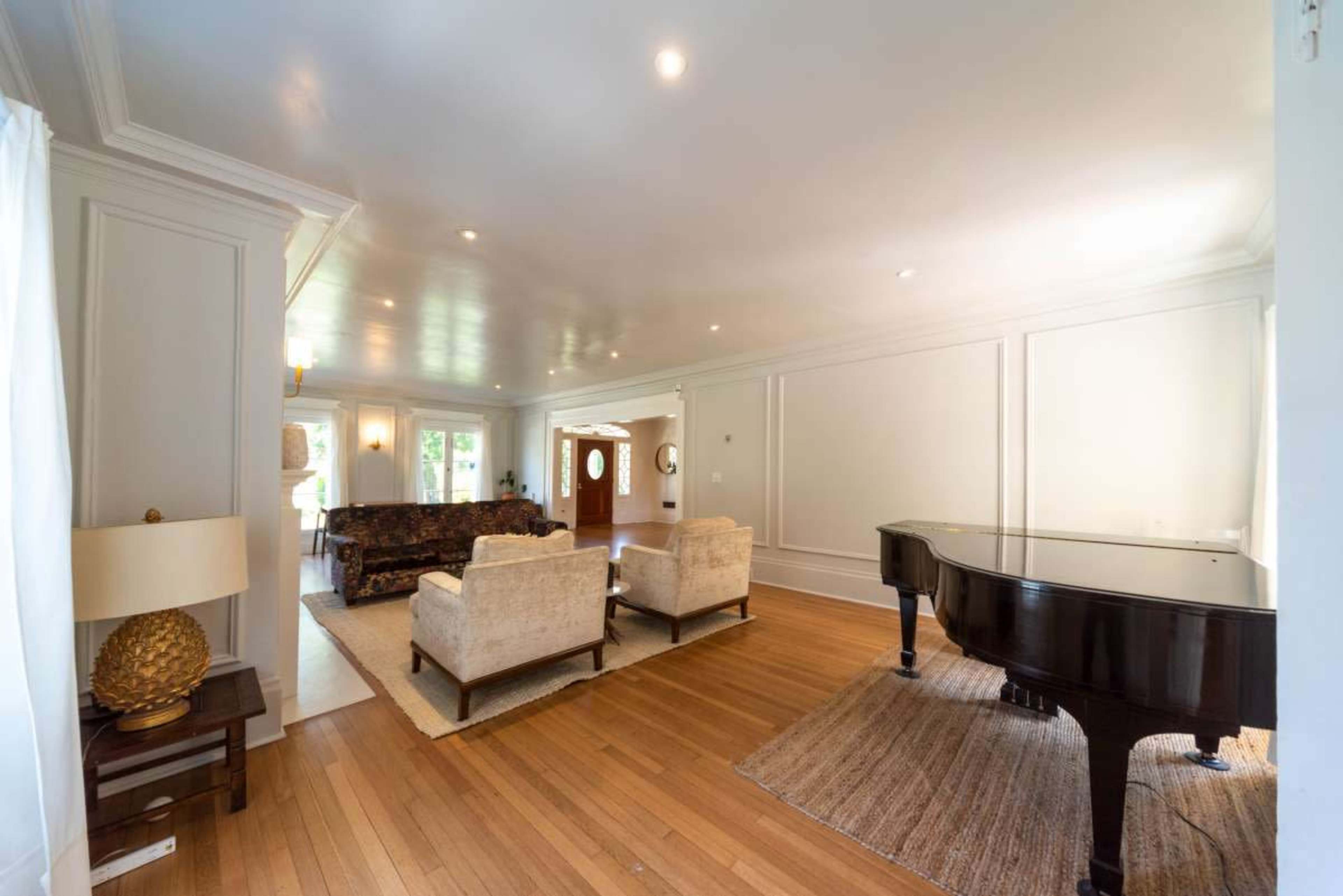 The image shows a spacious living room with a grand piano and two white armchairs arranged on wooden flooring near a patterned rug.