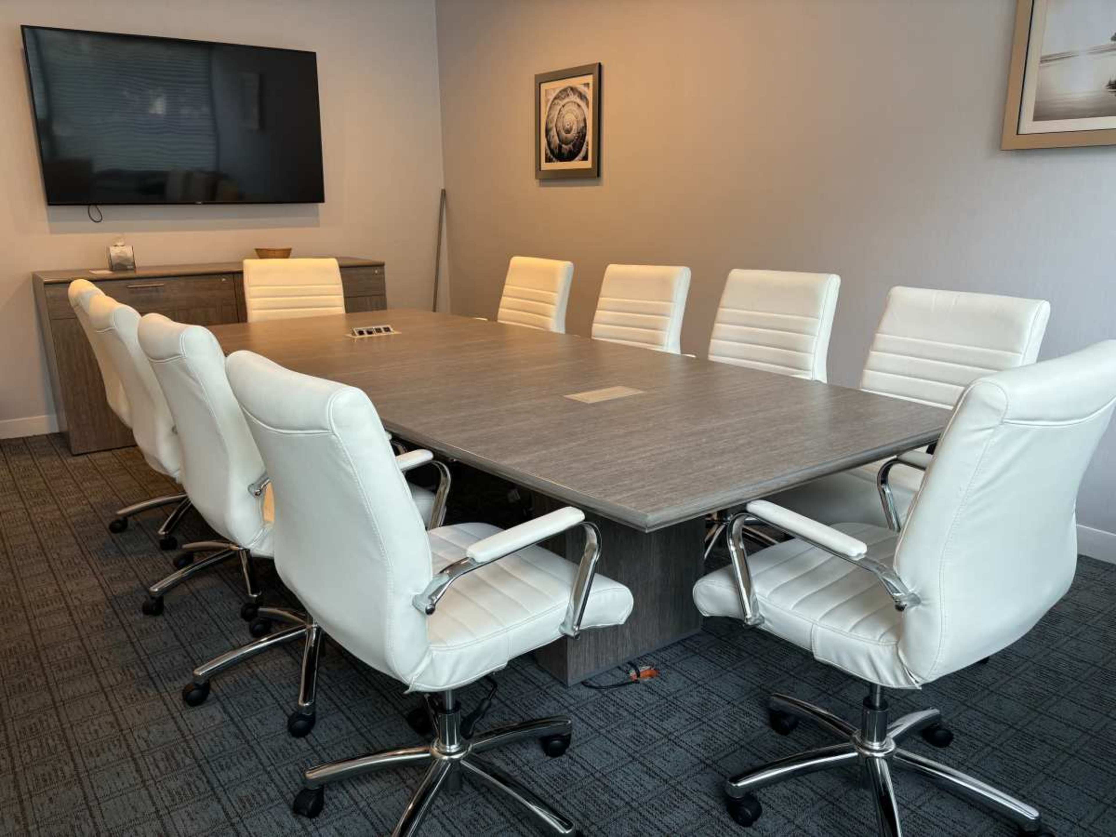 The image shows a modern conference room with a large rectangular table surrounded by eight white ergonomic chairs, a television mounted on the wall, and a side cabinet.