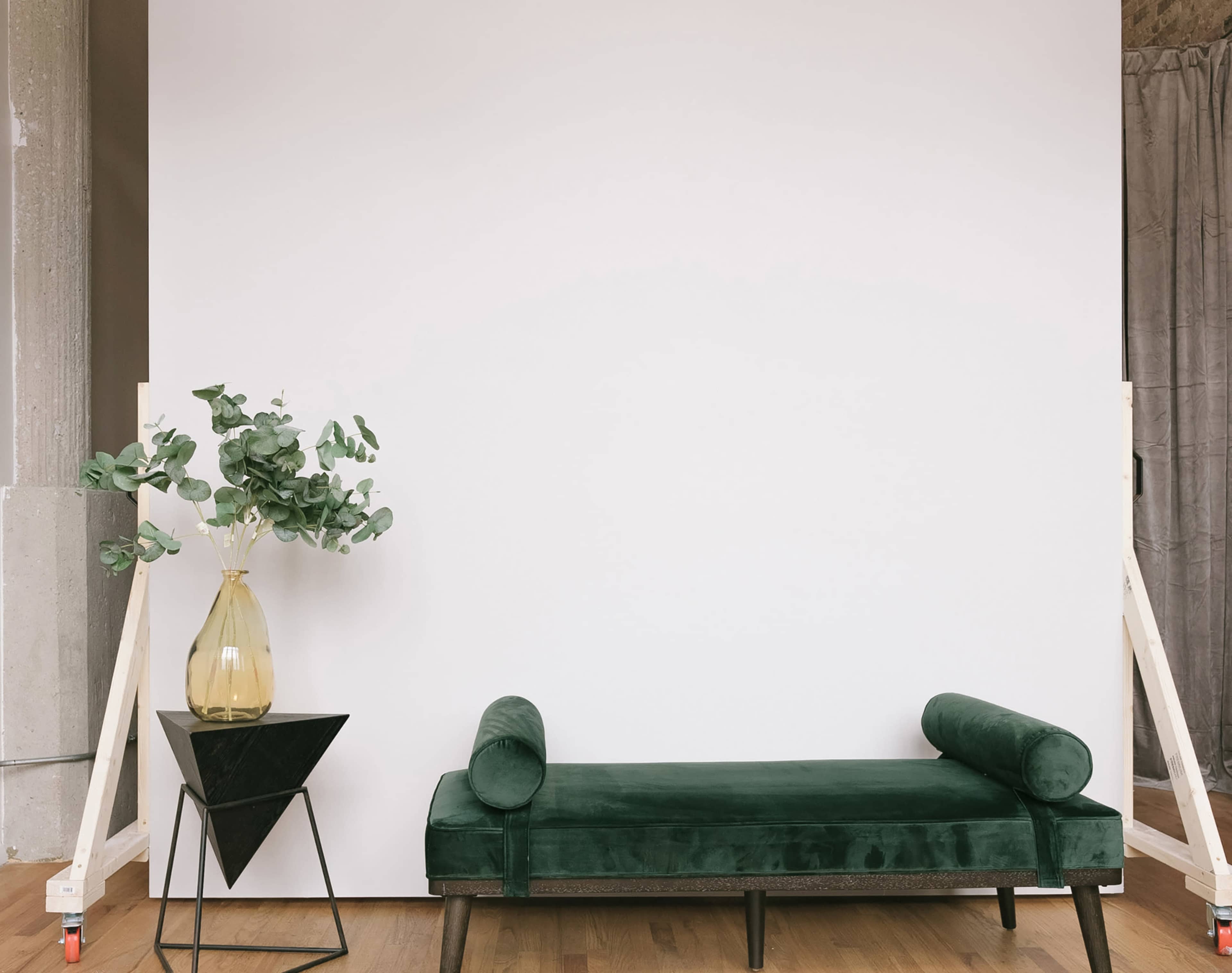 A green velvet bench is placed in front of a white wall, accompanied by a black triangular side table and a vase with greenery.
