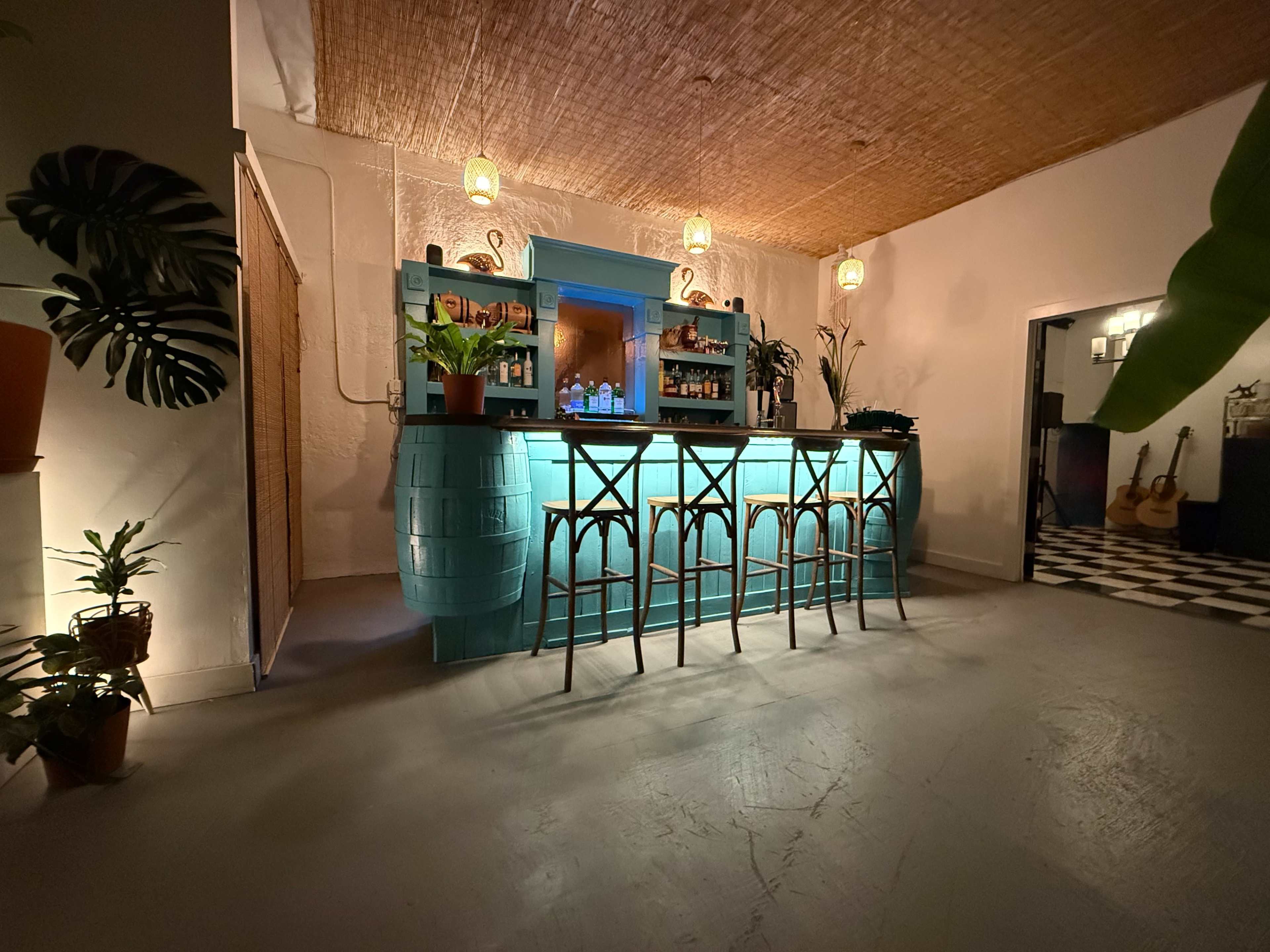 The image shows a brightly lit bar area with a turquoise bar counter, high stools, and various plants, set in a spacious room with a woven ceiling.