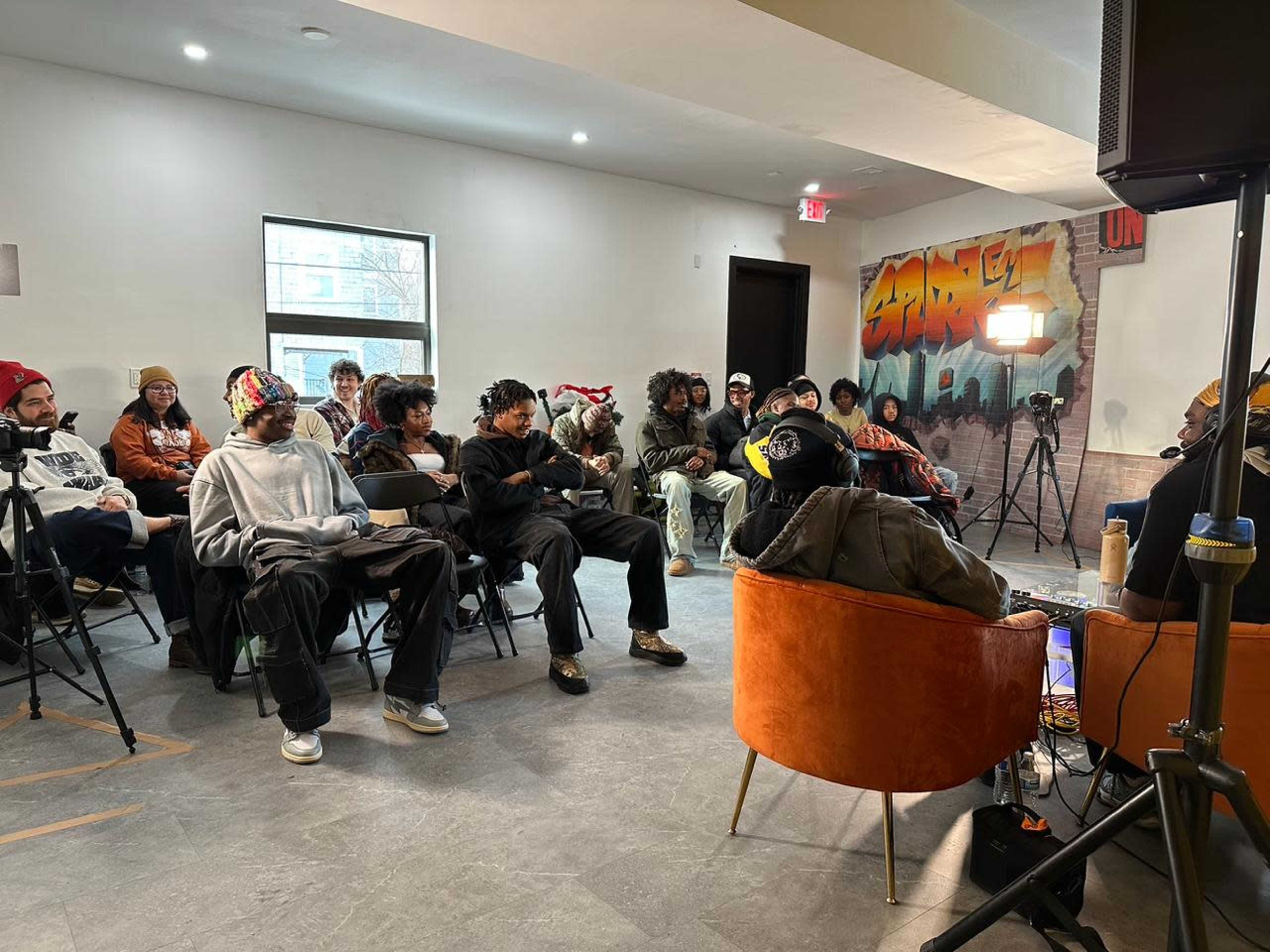 A group of people sits in chairs in a room with urban-themed artwork on the walls, facing a speaker.