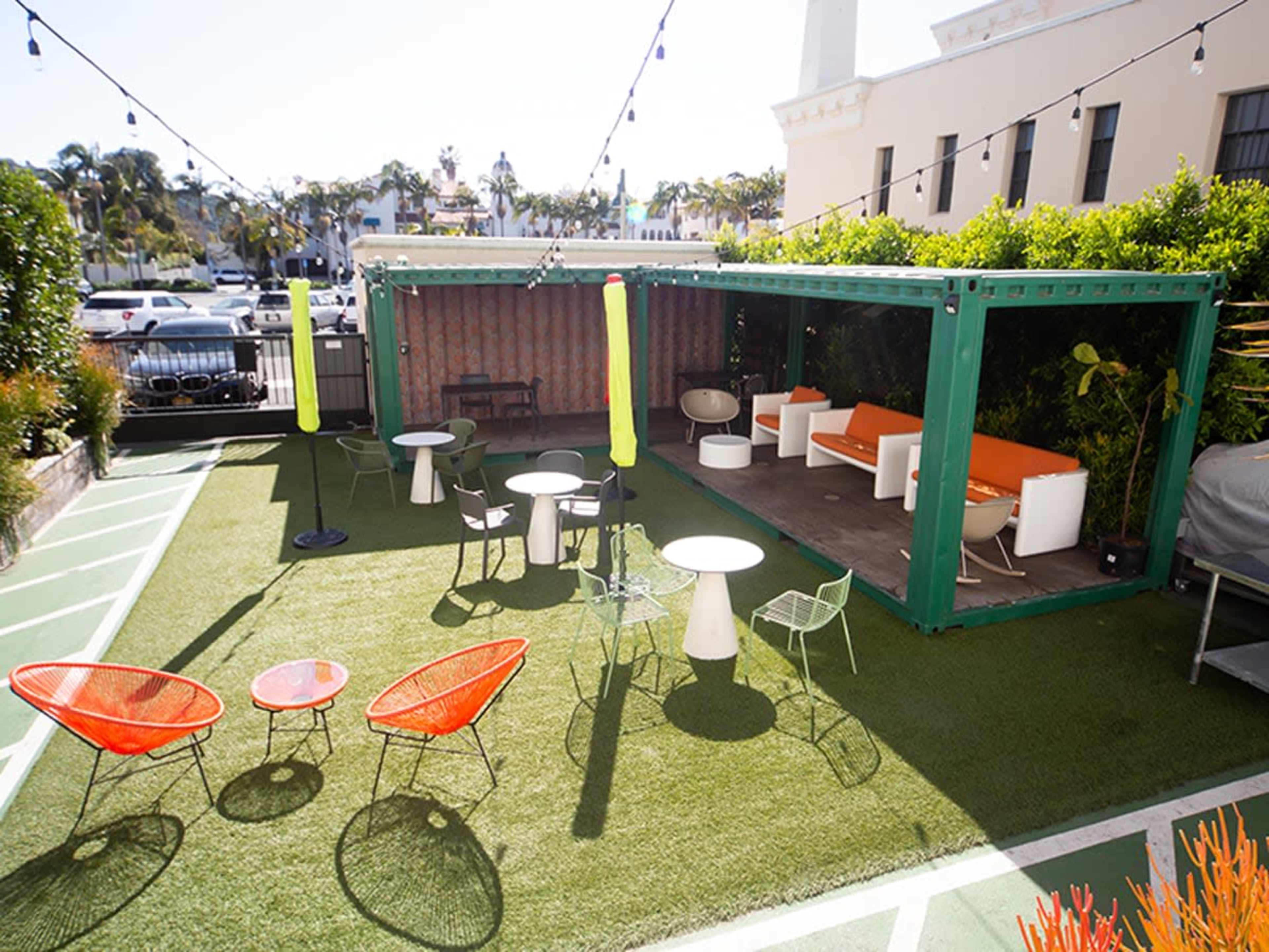 The image shows a modern outdoor seating area featuring green container-like structures, orange and white furniture, and several tables arranged on artificial grass.