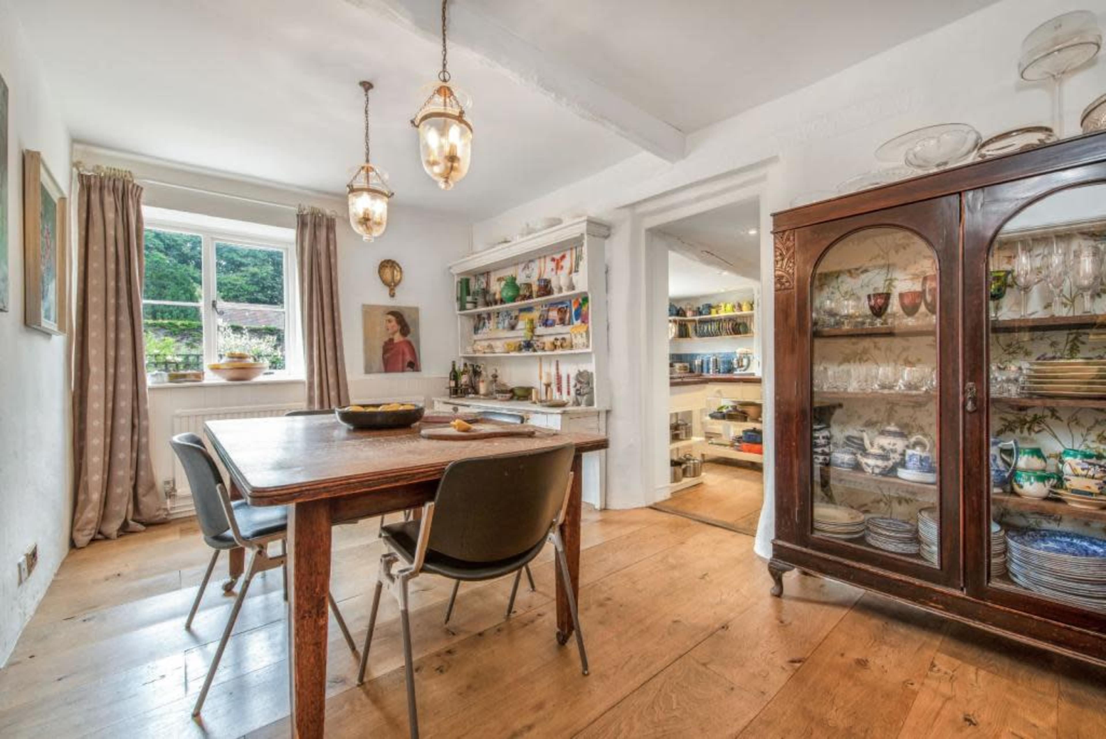 A wooden dining table with three chairs is situated in a well-lit room featuring a glass display cabinet and shelves filled with colorful dishes and artwork.