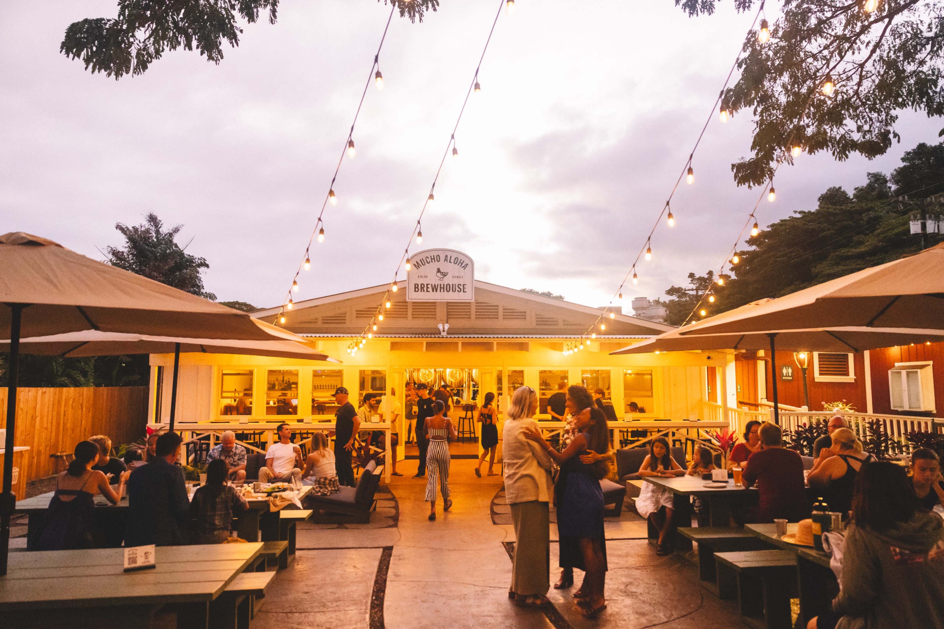 A bustling outdoor brewhouse features patrons dining and socializing under string lights as twilight approaches.