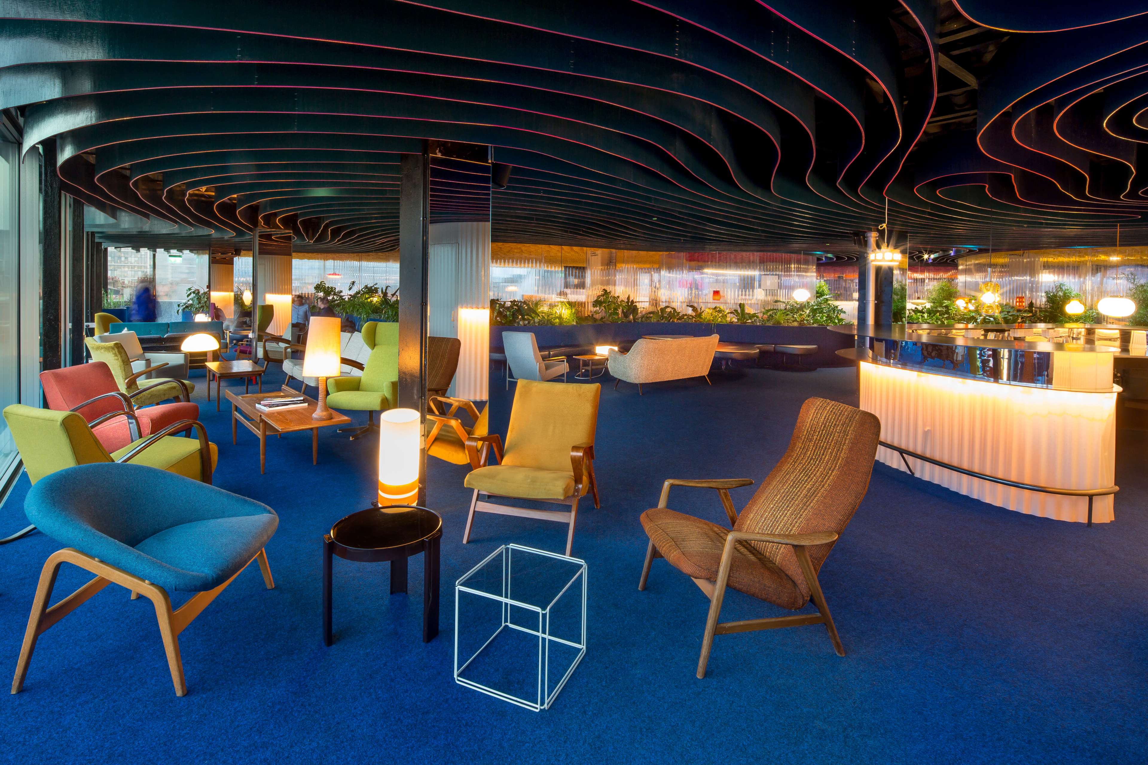 The image features a modern lounge area with colorful chairs, a blue carpet, and a softly lit bar in the background.