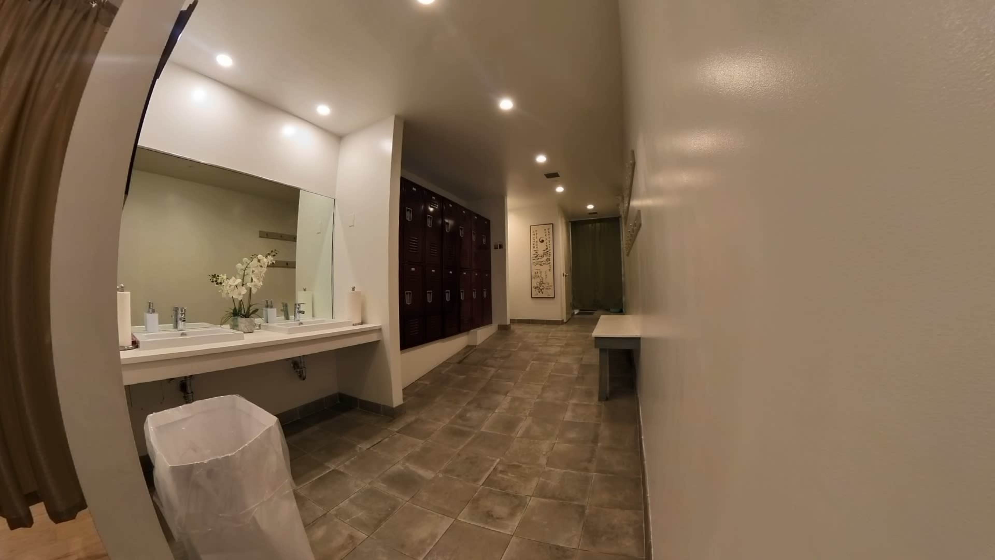 The image shows a spacious restroom with a long mirror, sink area, and rows of lockers along the side walls.