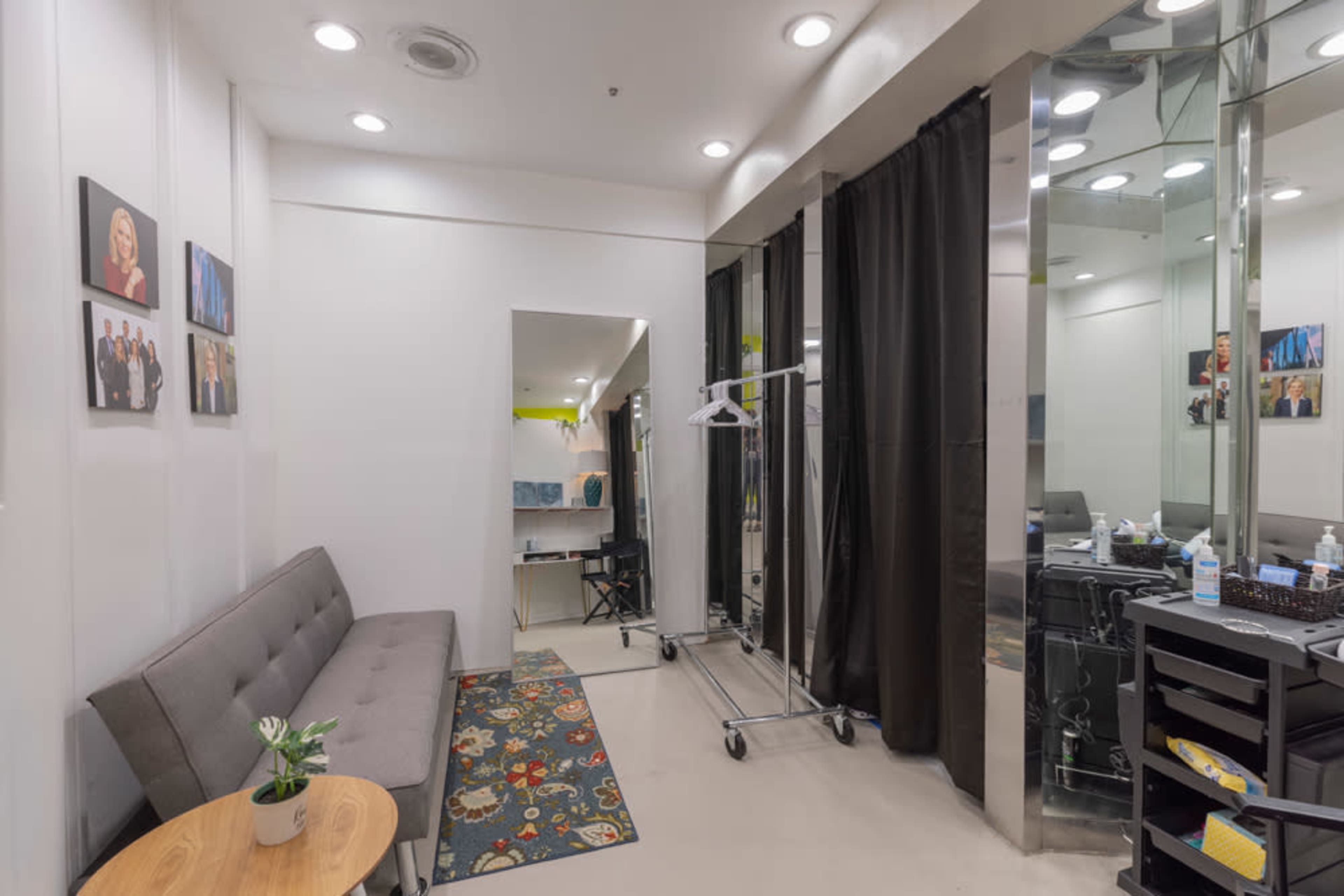 A small, modern waiting room features a gray sofa, a round wooden table, and a rack with hangers, with a mirrored wall and several framed photographs.