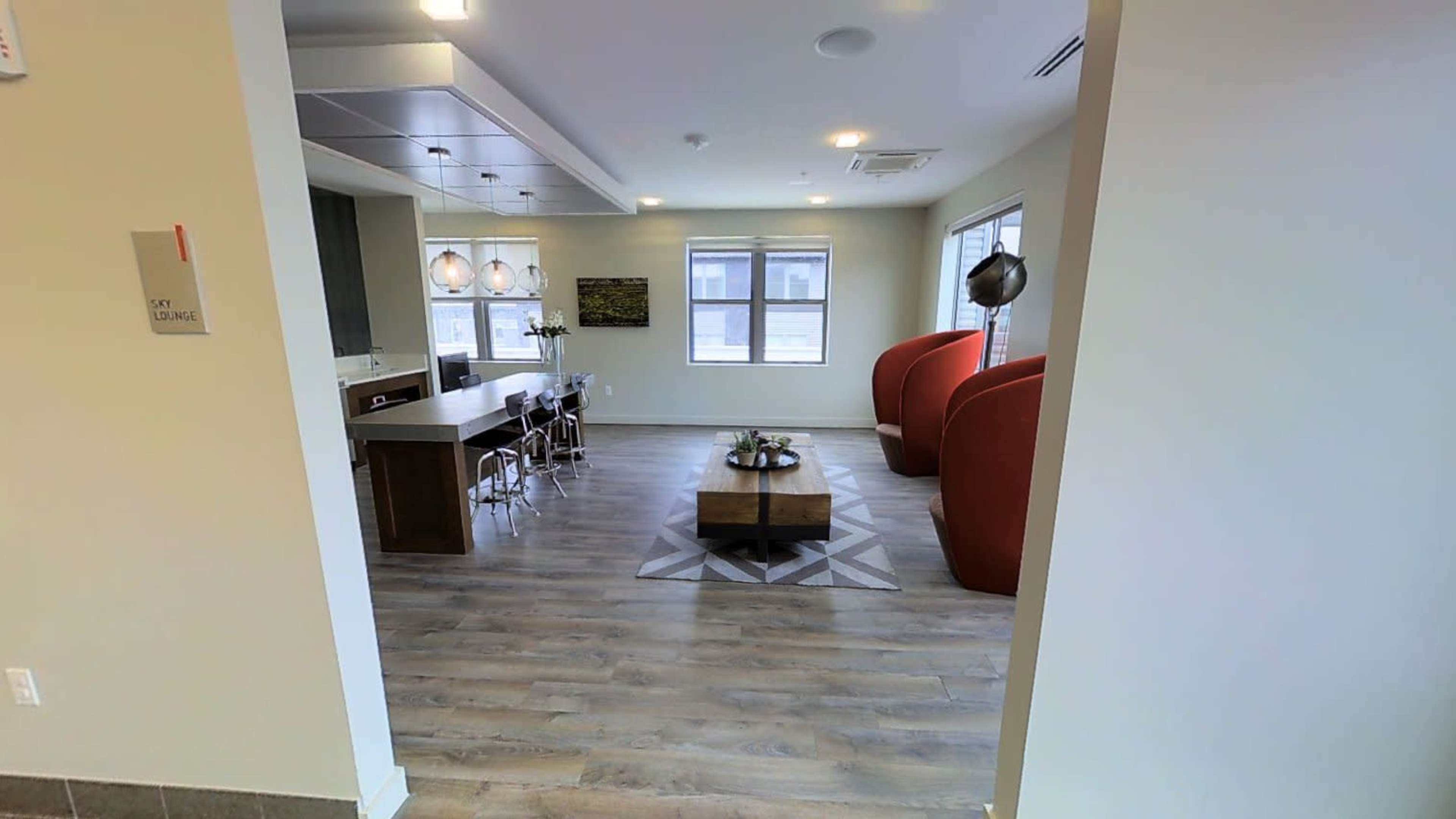 A modern lounge area featuring a kitchen space, two red accent chairs, and a coffee table on a patterned rug.