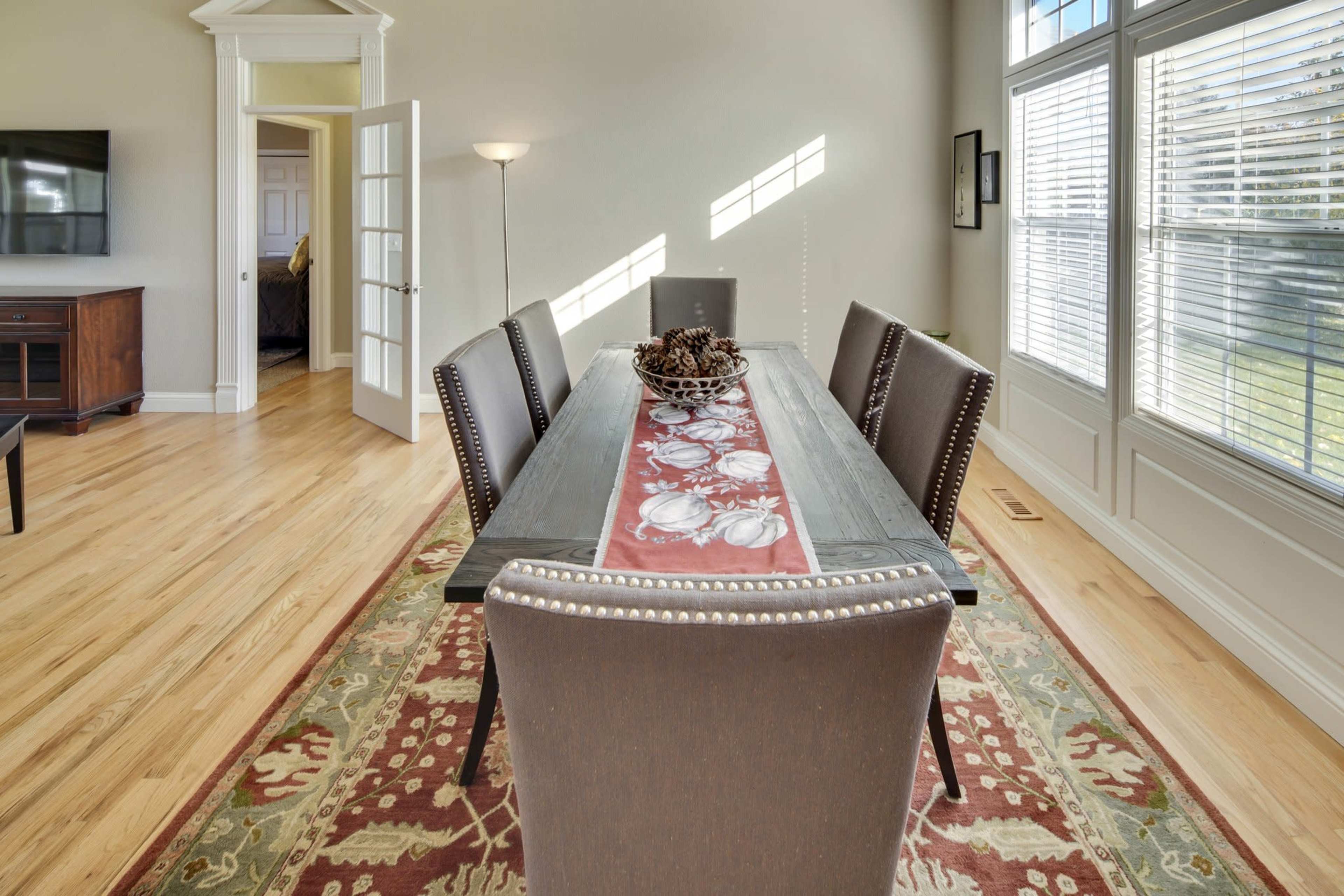 A dining table with six chairs is set in a well-lit room, featuring large windows and hardwood floors, with a decorative centerpiece on the table.