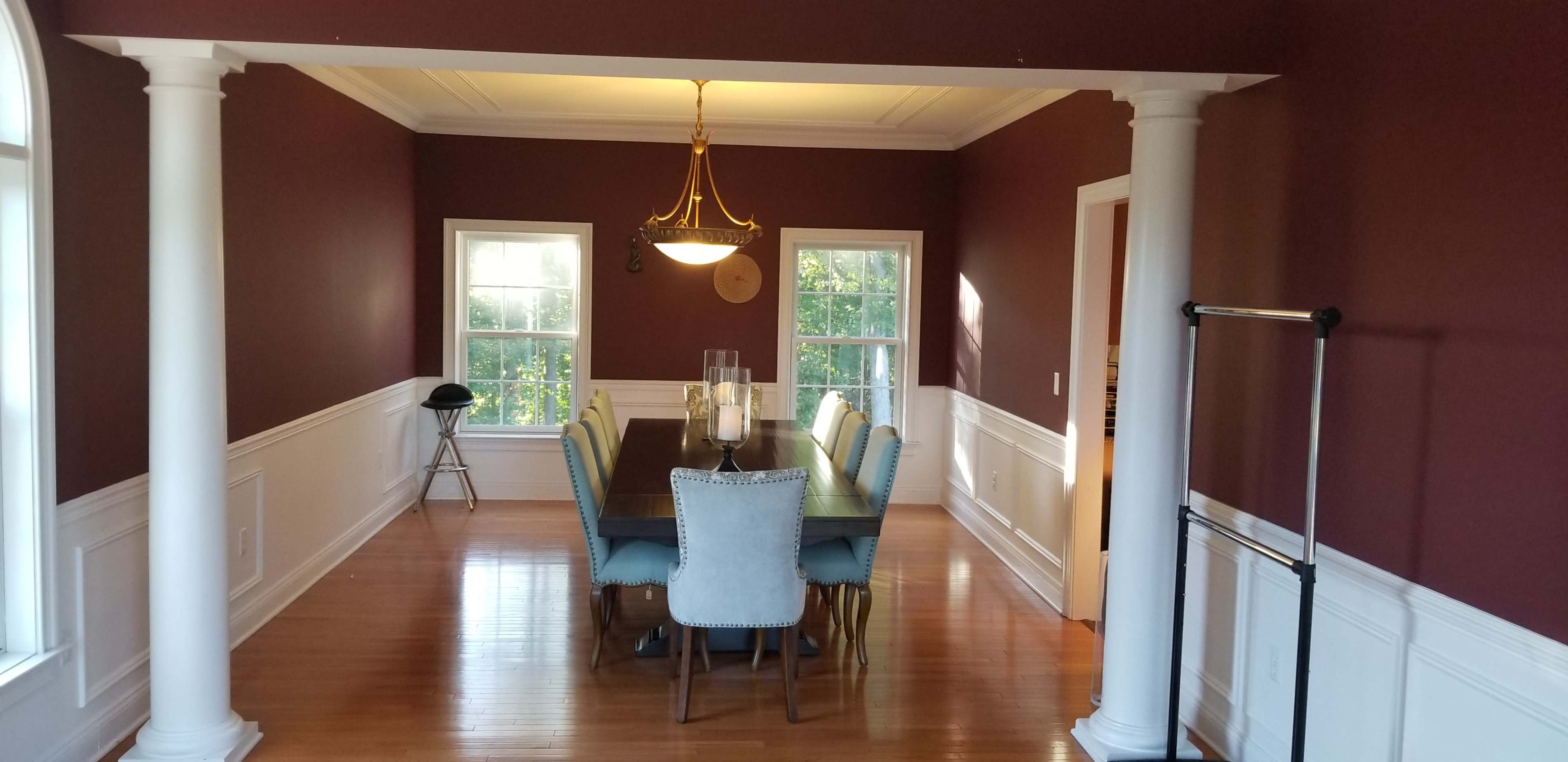 The image shows a dining room with a long table surrounded by upholstered chairs, flanked by two windows and decorated with a chandelier.