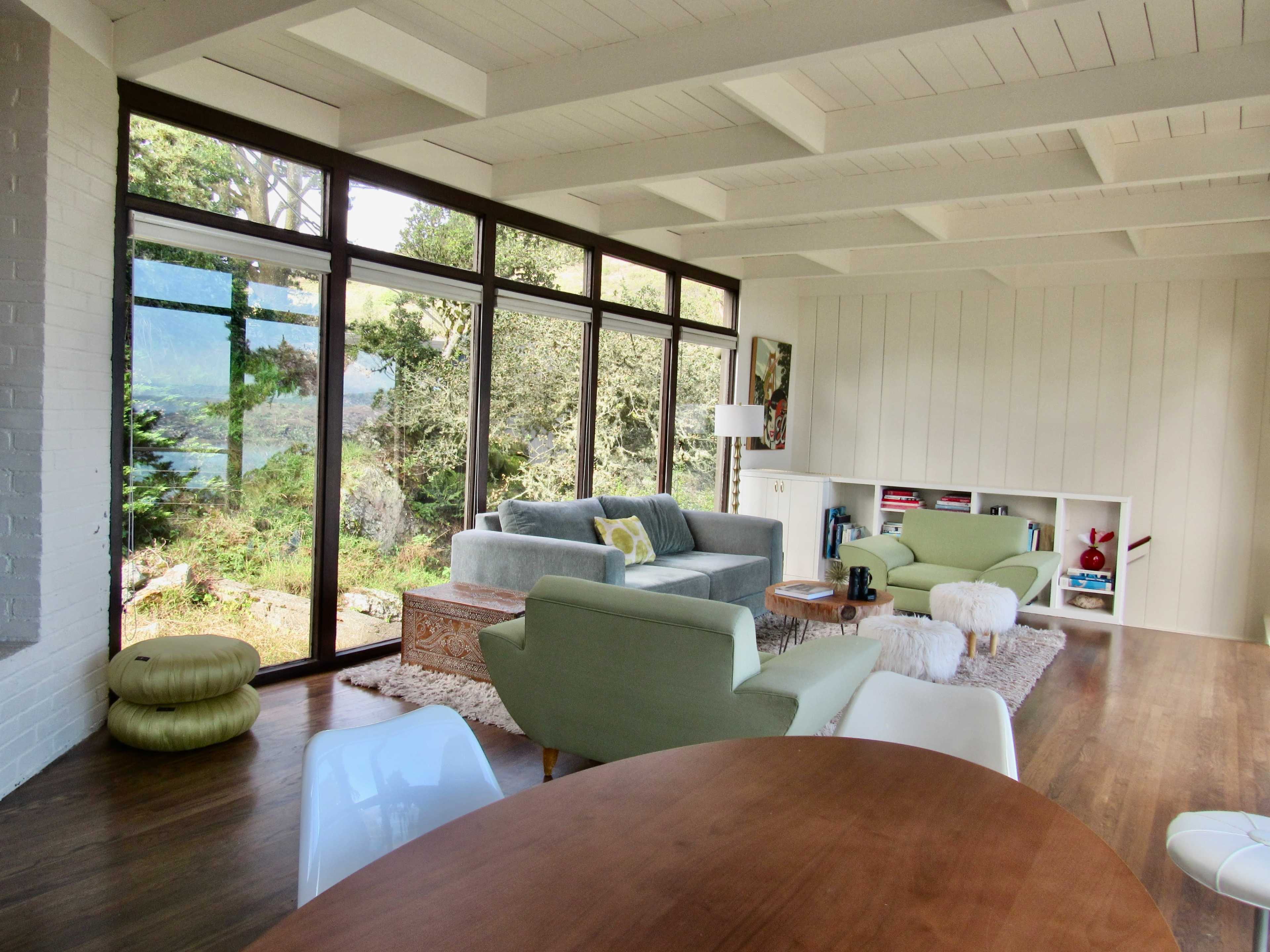 A modern living room with large windows overlooking a natural landscape, featuring a gray sofa, green chairs, and a wooden dining table.