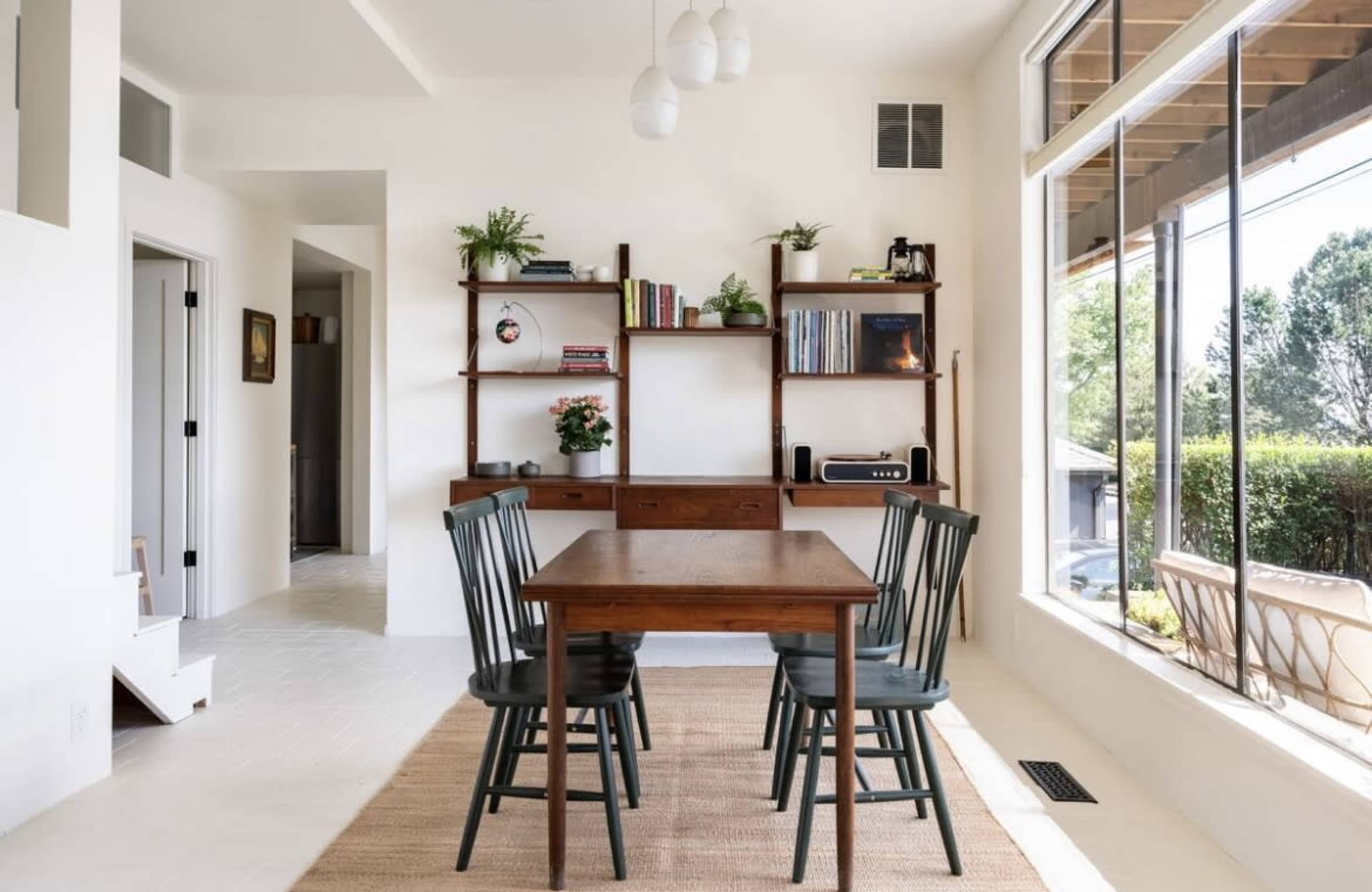 A wooden dining table with four green chairs is positioned in a bright room featuring large windows, a bookshelf, and a jute rug.