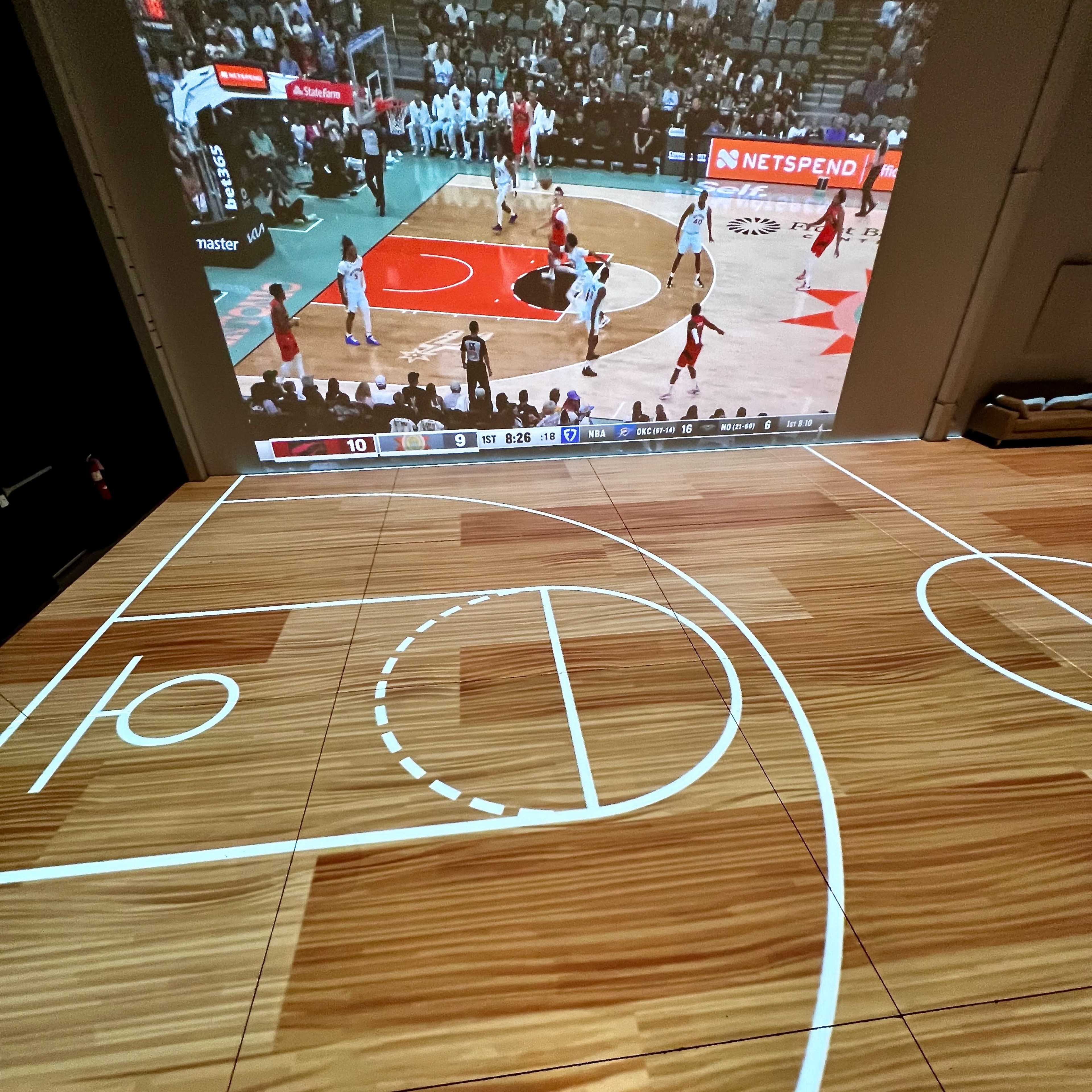 A basketball court is projected onto a floor while a game is displayed on a screen in the background.
