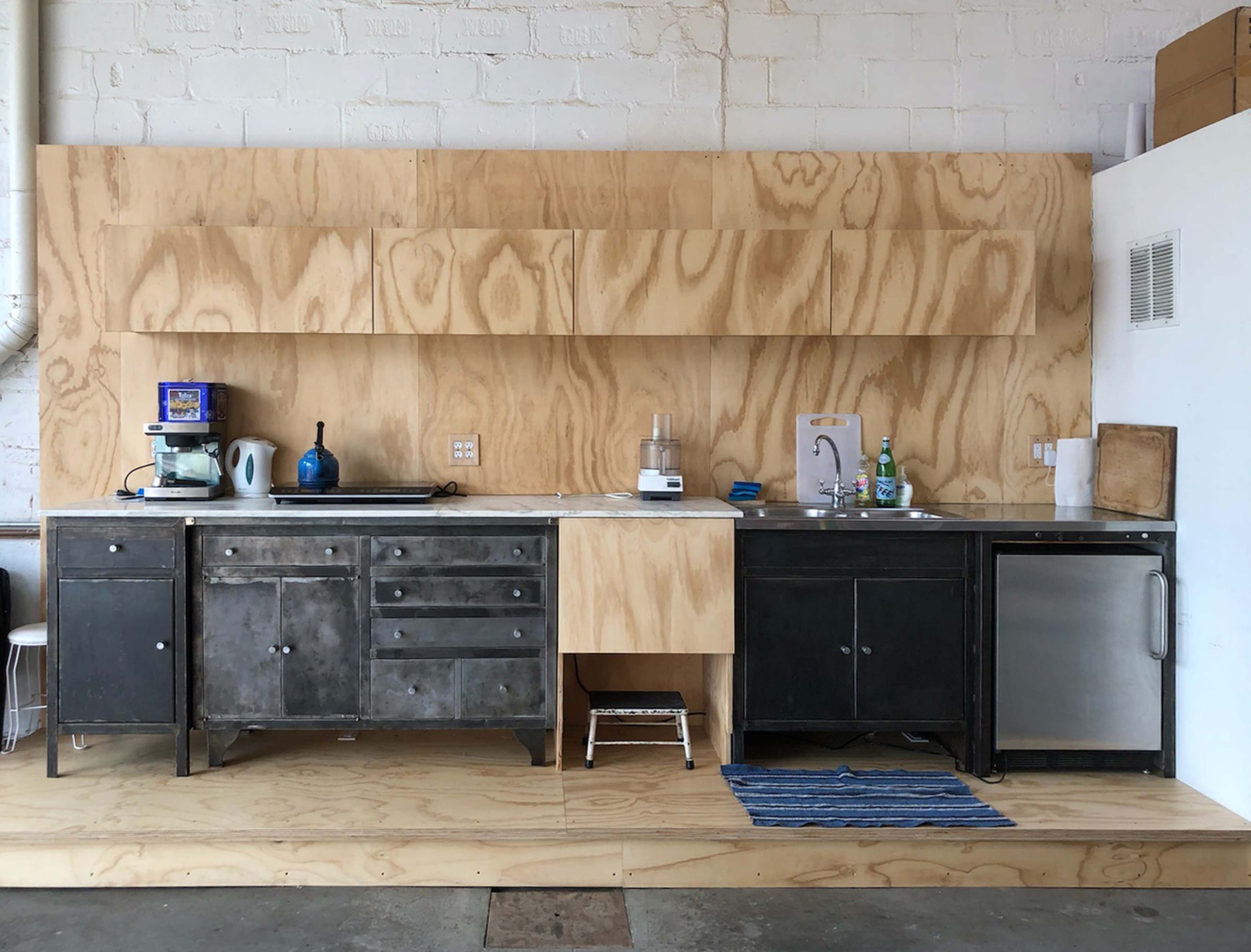 The image shows a minimalist kitchen with a wooden wall, metal cabinets, a sink, and a stainless steel refrigerator.
