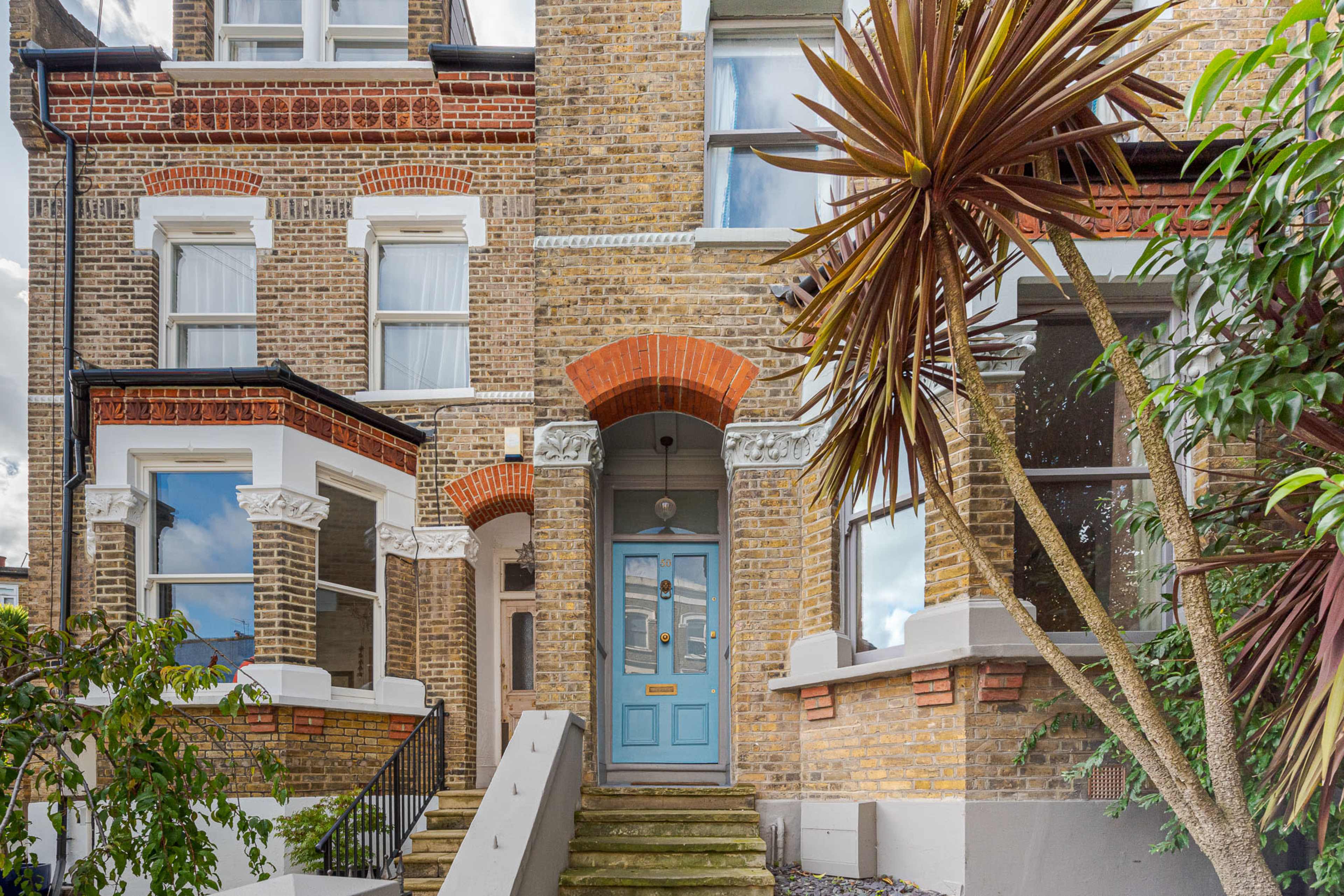 The image shows a brick townhouse featuring a blue front door, surrounded by greenery and decorative plants.