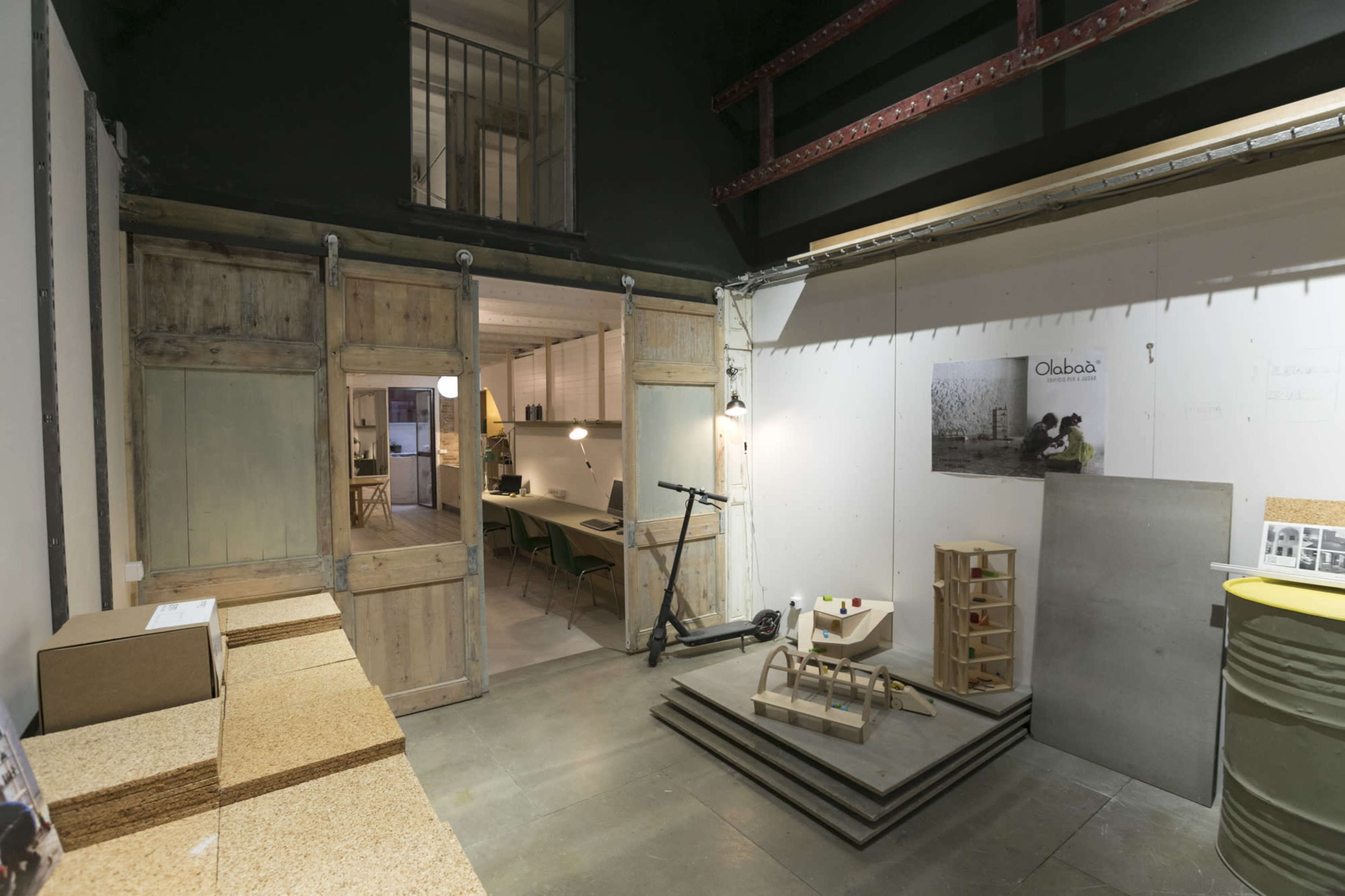 An interior space featuring wooden sliding doors, a scooter, and stacks of cork boards with tables and chairs in the background.