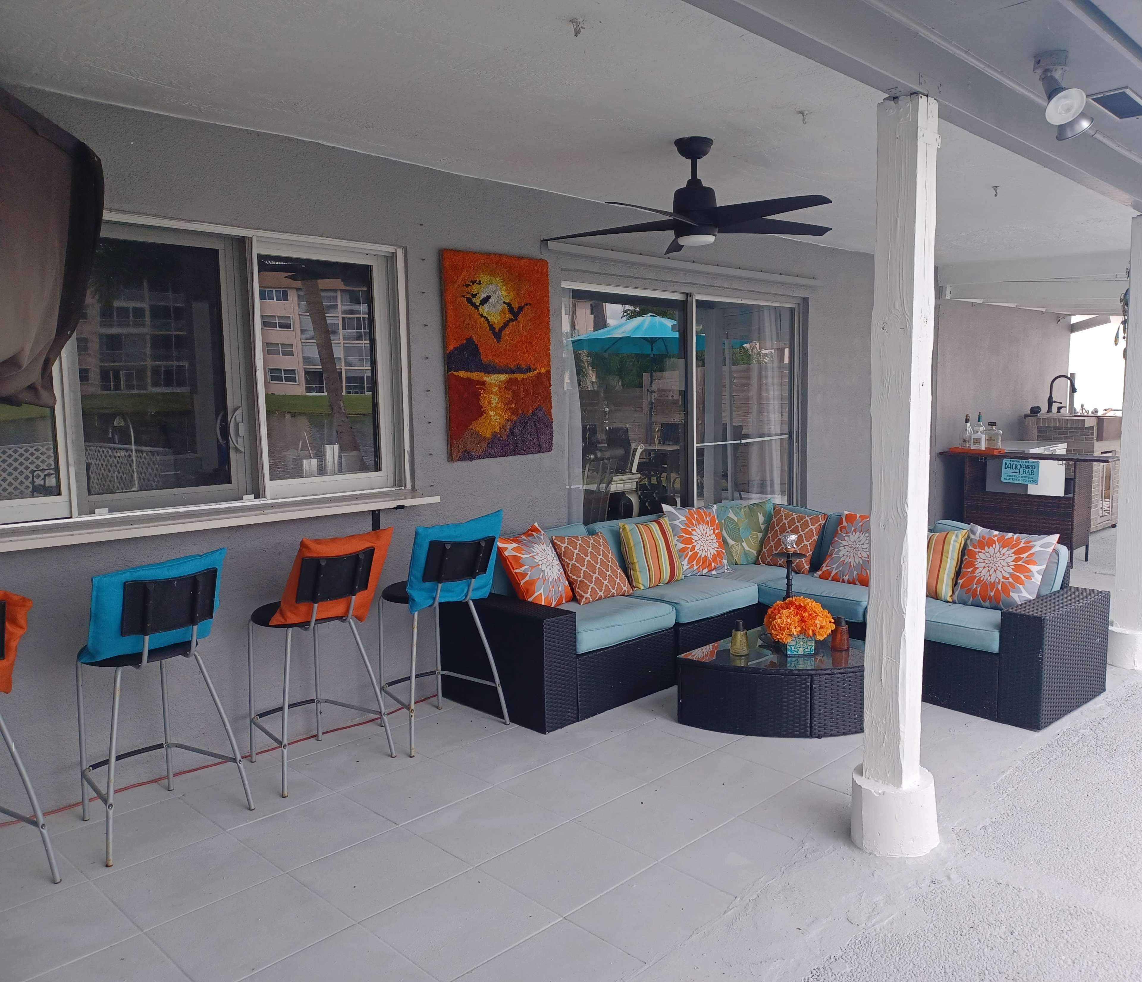 A covered patio features a wicker seating area adorned with colorful cushions, alongside a bar with stools and a backdrop of a beachside view.