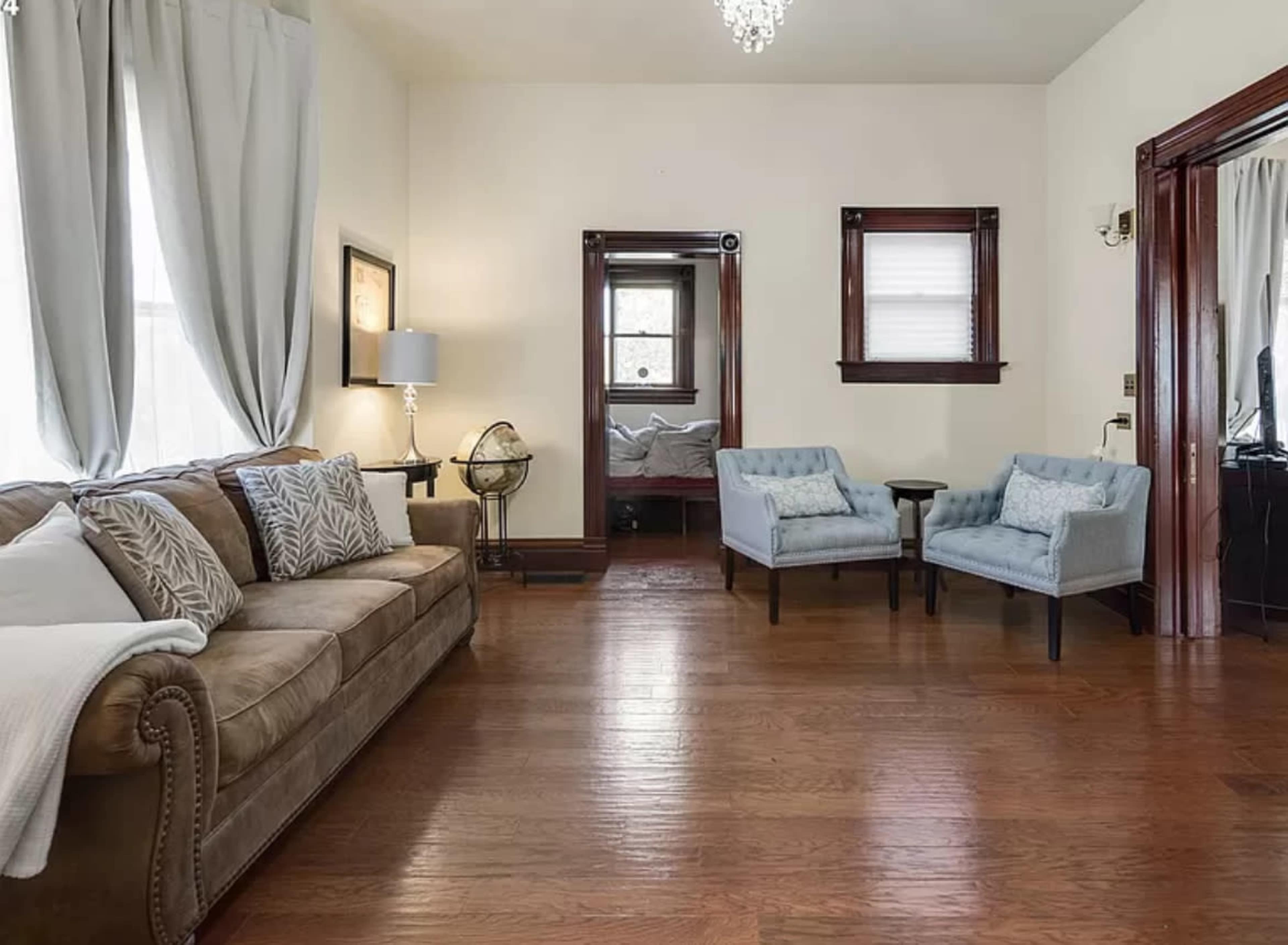 A living room with a brown sofa, two blue chairs, and wooden flooring, along with a small round table and a doorway leading to another room.