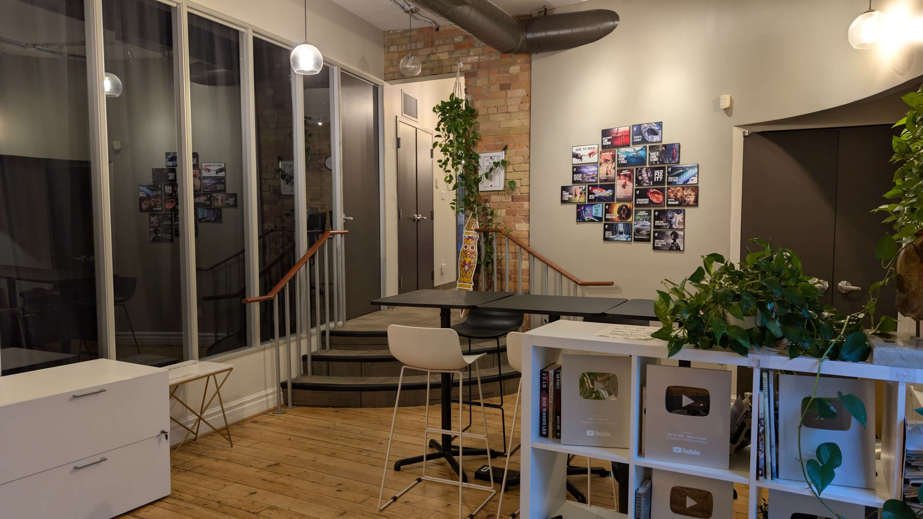 The image shows a modern interior space with wooden floors, a staircase leading to another level, a table and chairs, and a wall adorned with various framed photographs.