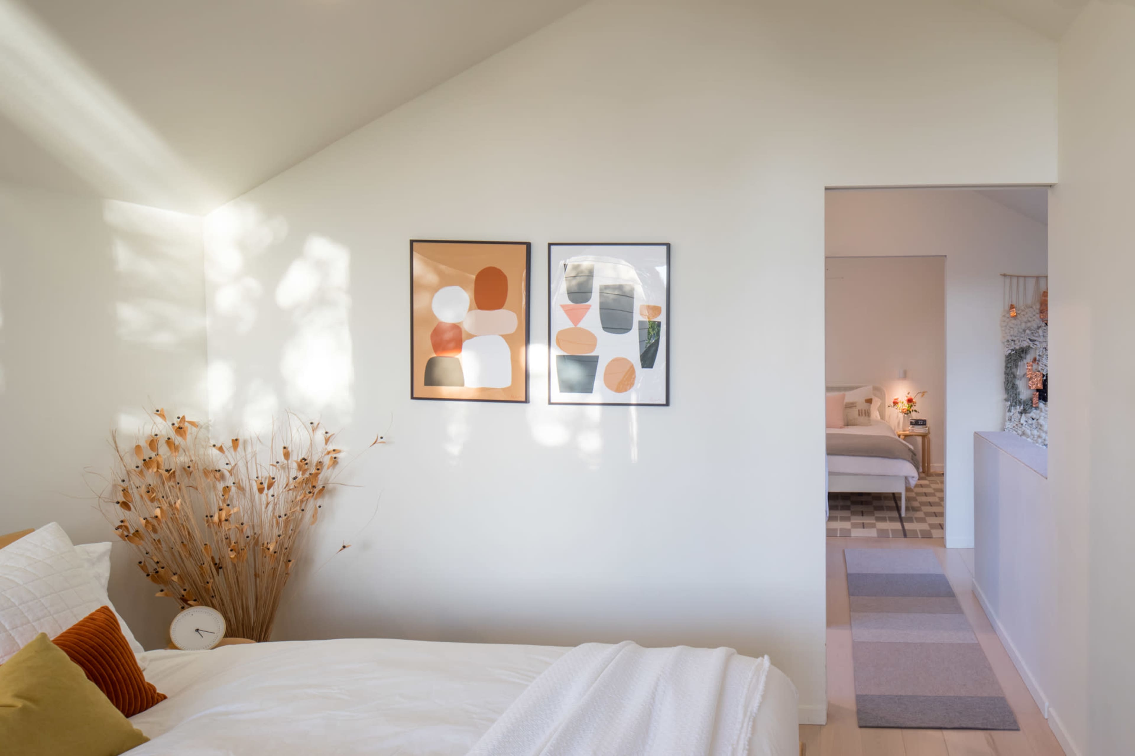 A bright bedroom features a large bed with cozy bedding, two framed abstract prints on the wall, and a view of another room through an open doorway.