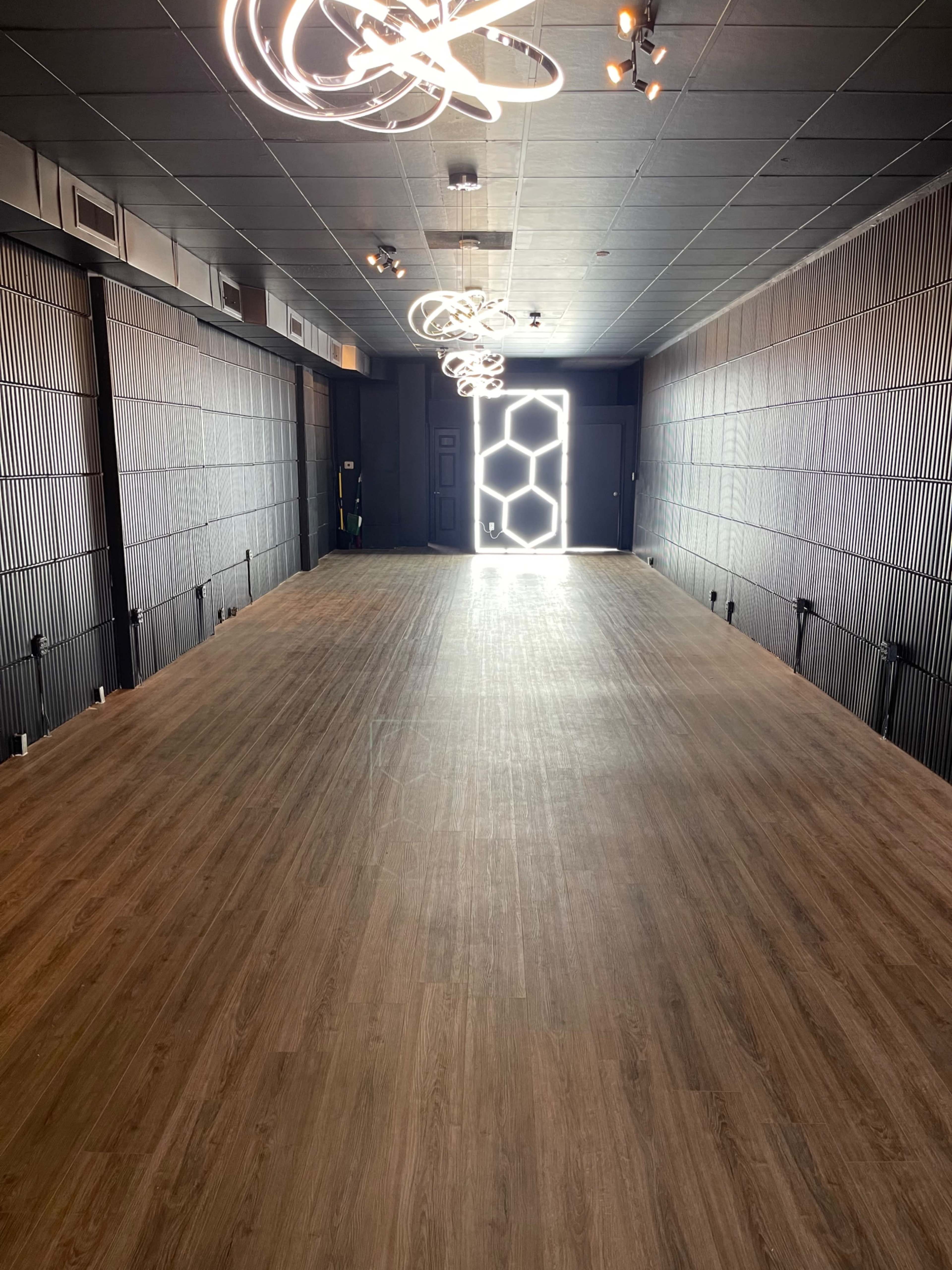 A spacious, well-lit room with wooden flooring and modern light fixtures, leading to a door at the far end.