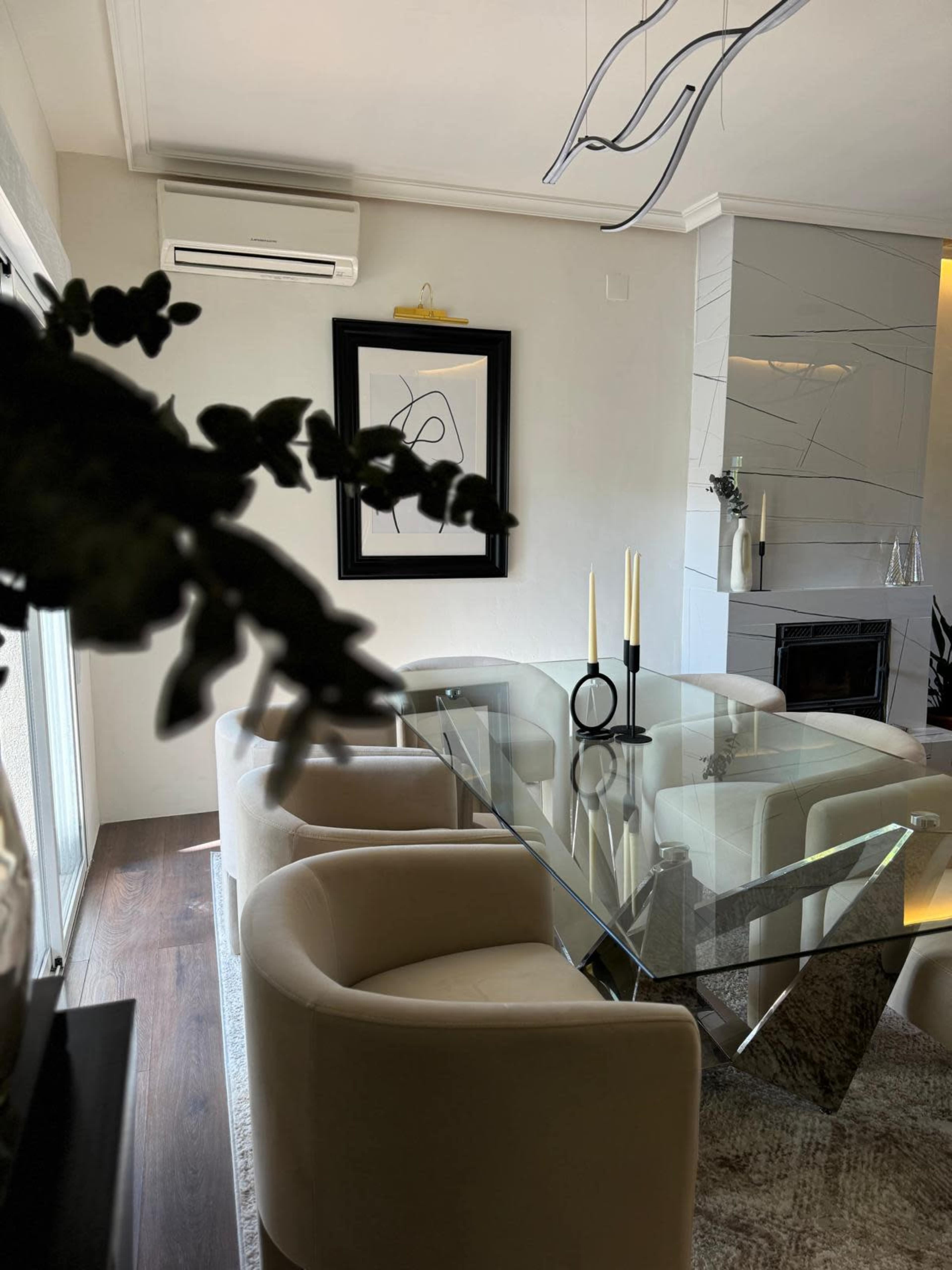 A modern dining area features a glass table surrounded by cream-colored chairs, with a black and white artwork on the wall and candles on the table.