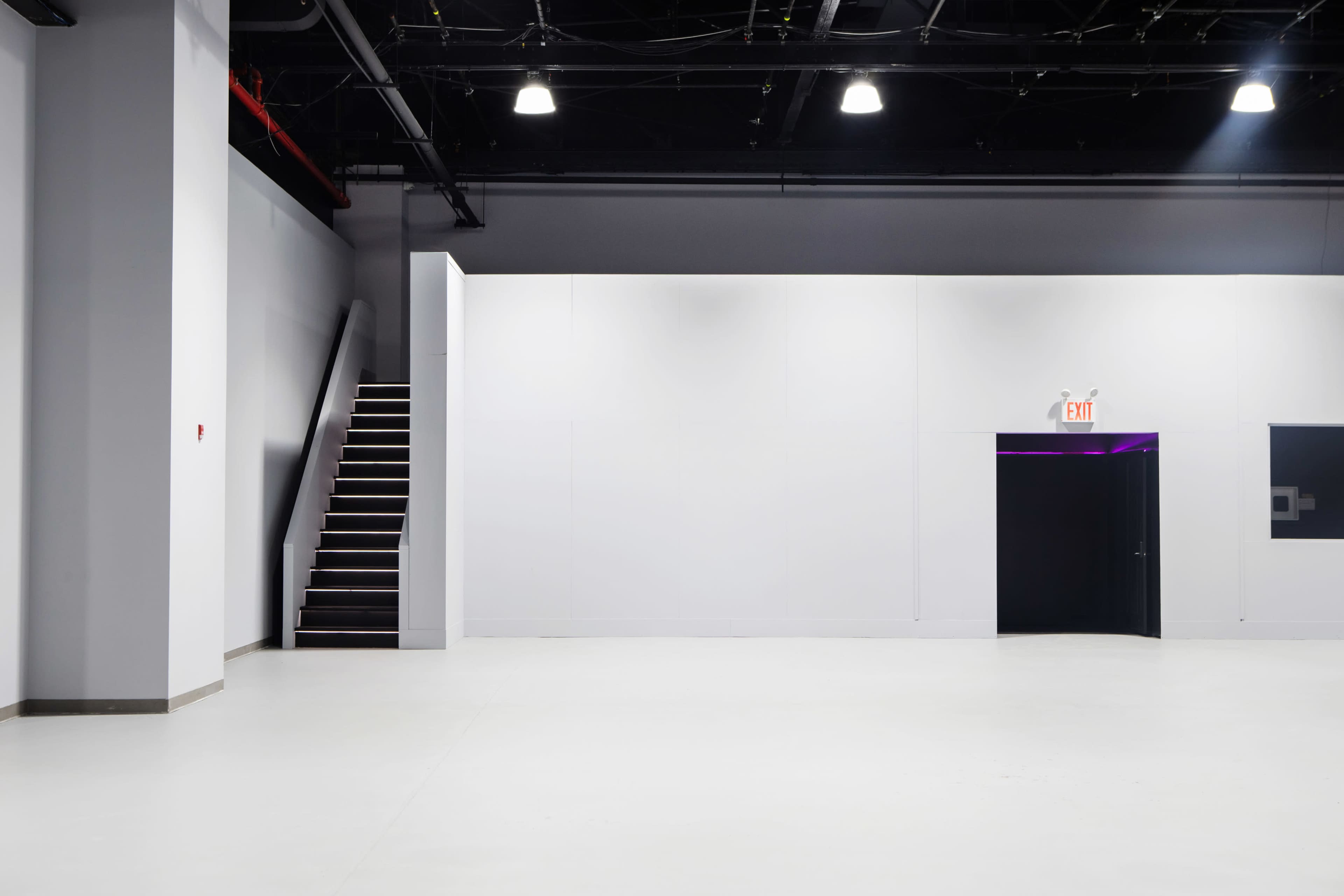 The image shows a spacious, empty interior with white walls, a set of stairs on the left, and a black door on the right.