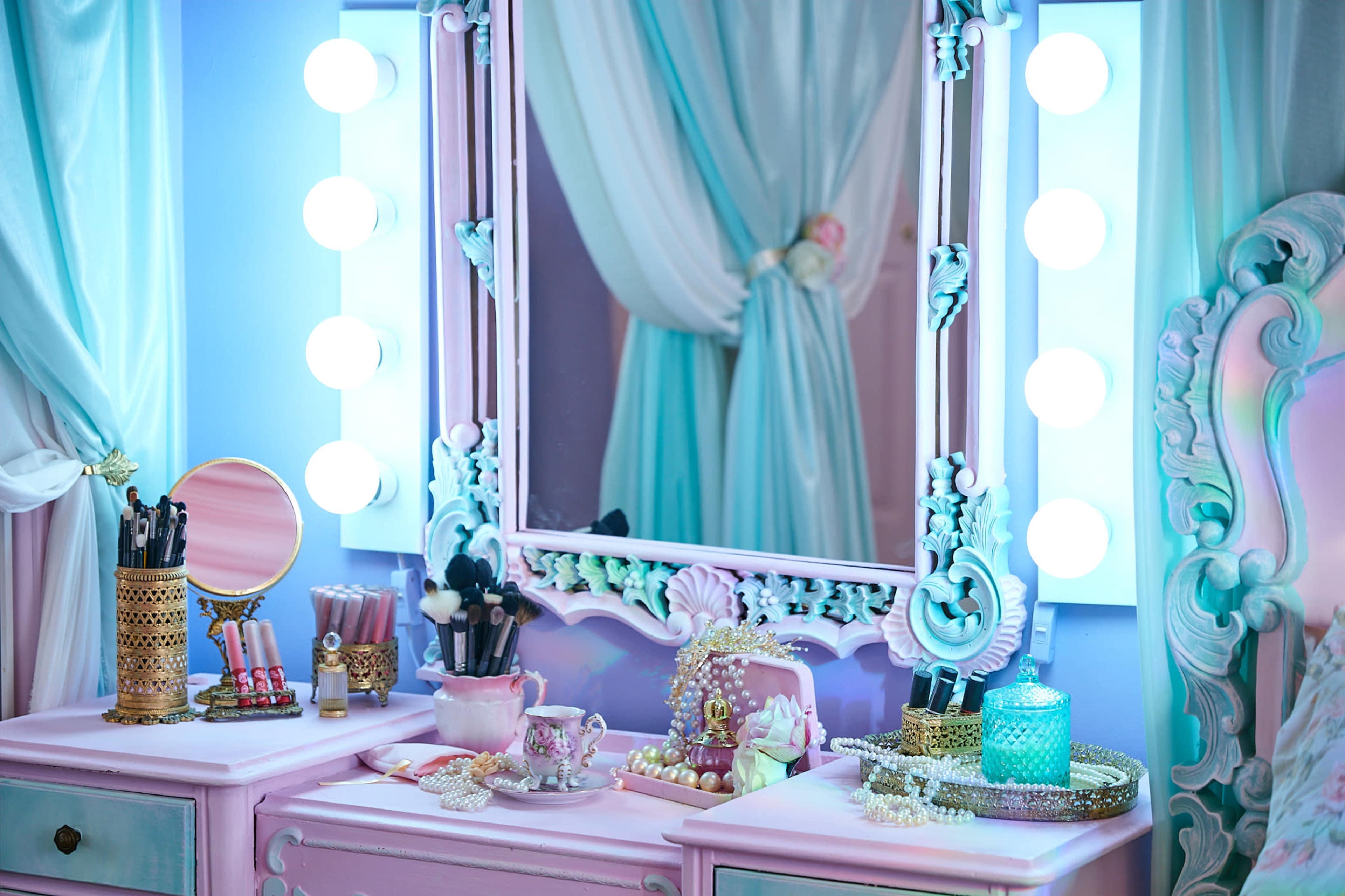 A vanity table is set against a softly lit backdrop, featuring a large mirror surrounded by illuminated bulbs, with various makeup tools and accessories neatly arranged on the surface.