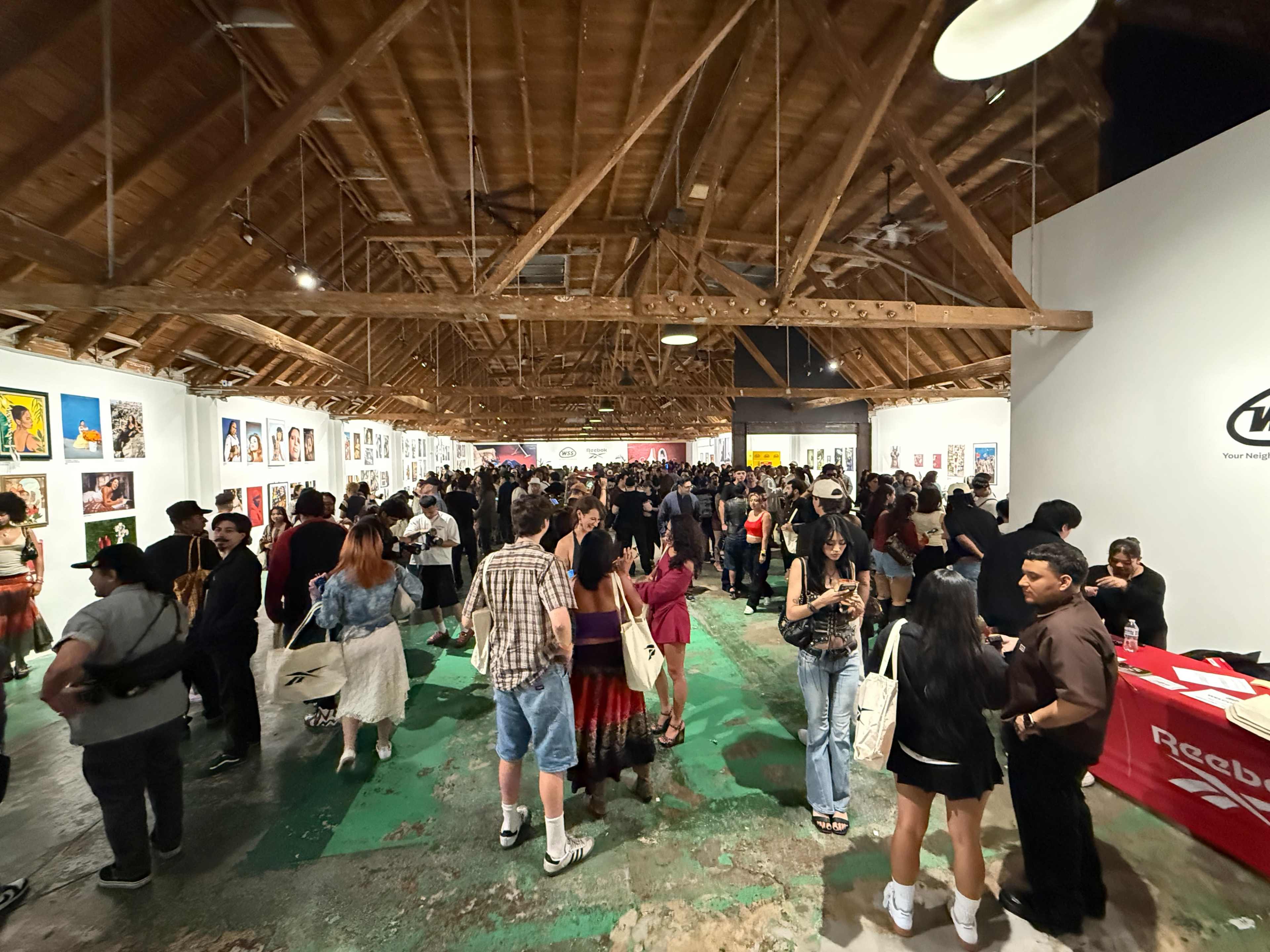 A large crowd gathers in an art gallery with wooden beams and artwork displayed on the walls.