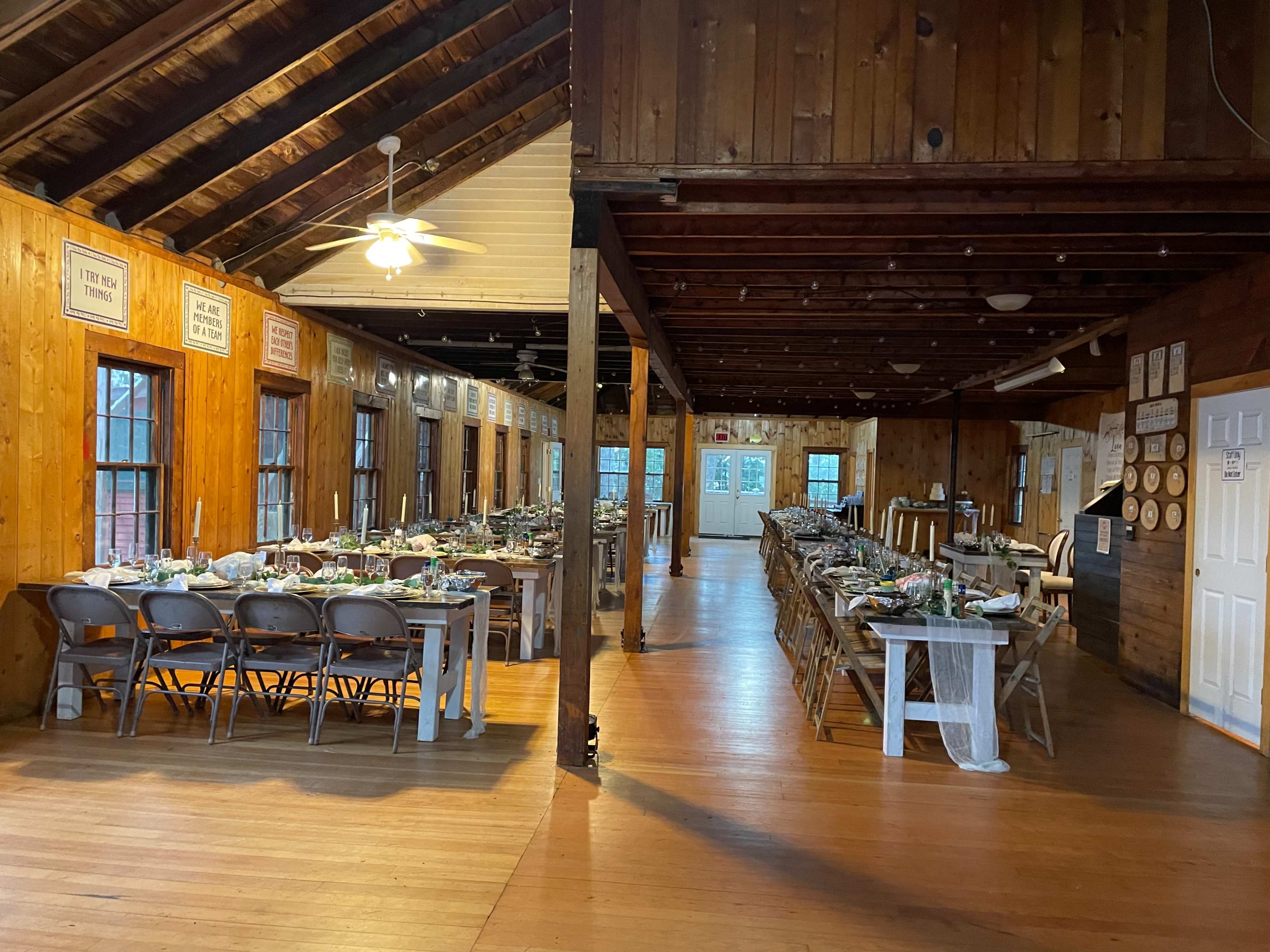 A wooden hall set up for a dining event, featuring neatly arranged tables with floral centerpieces and chairs, under a slatted wooden ceiling.