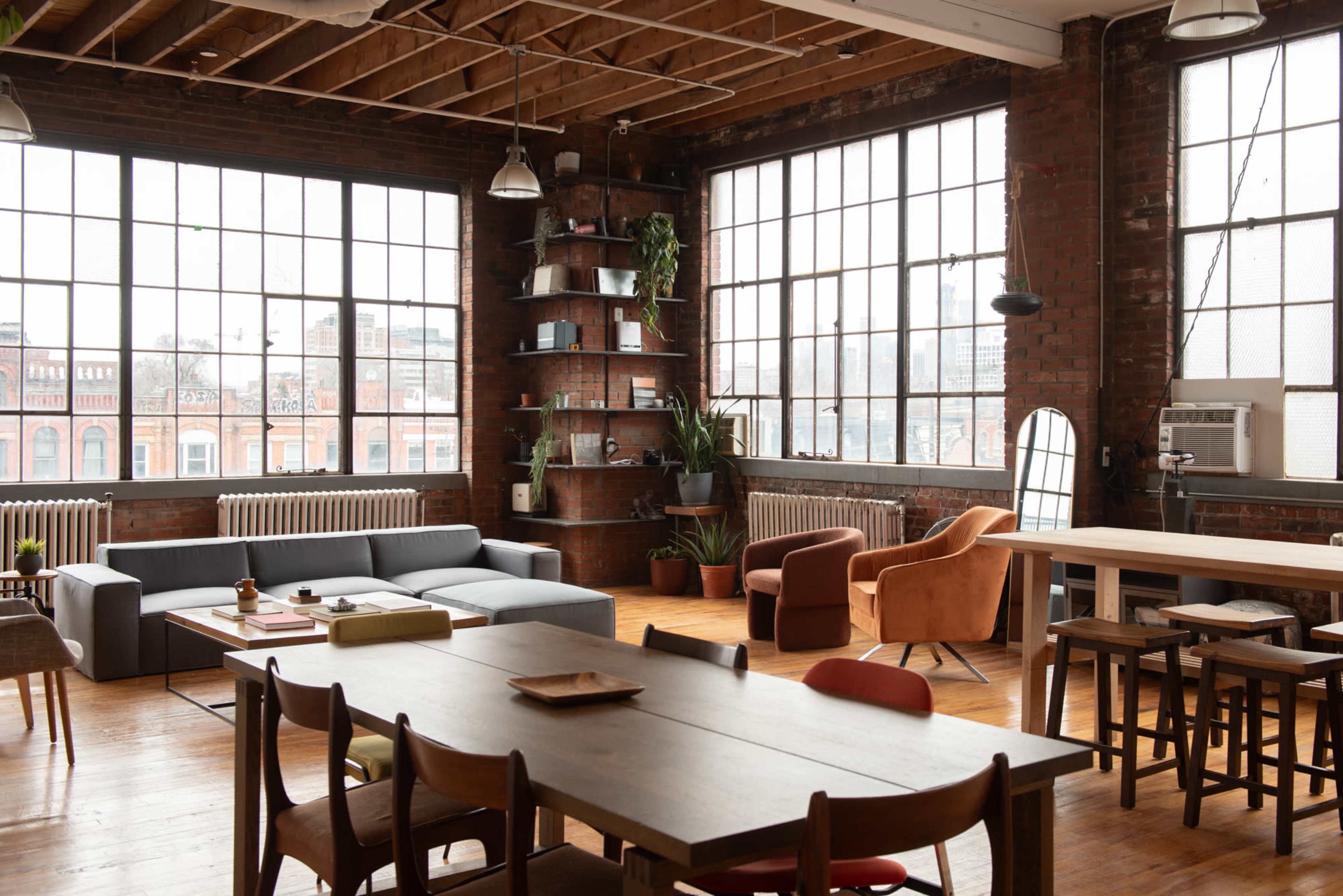 The image shows a spacious industrial-style living area with large windows, a mix of seating options, a wooden table, and shelves adorned with plants.